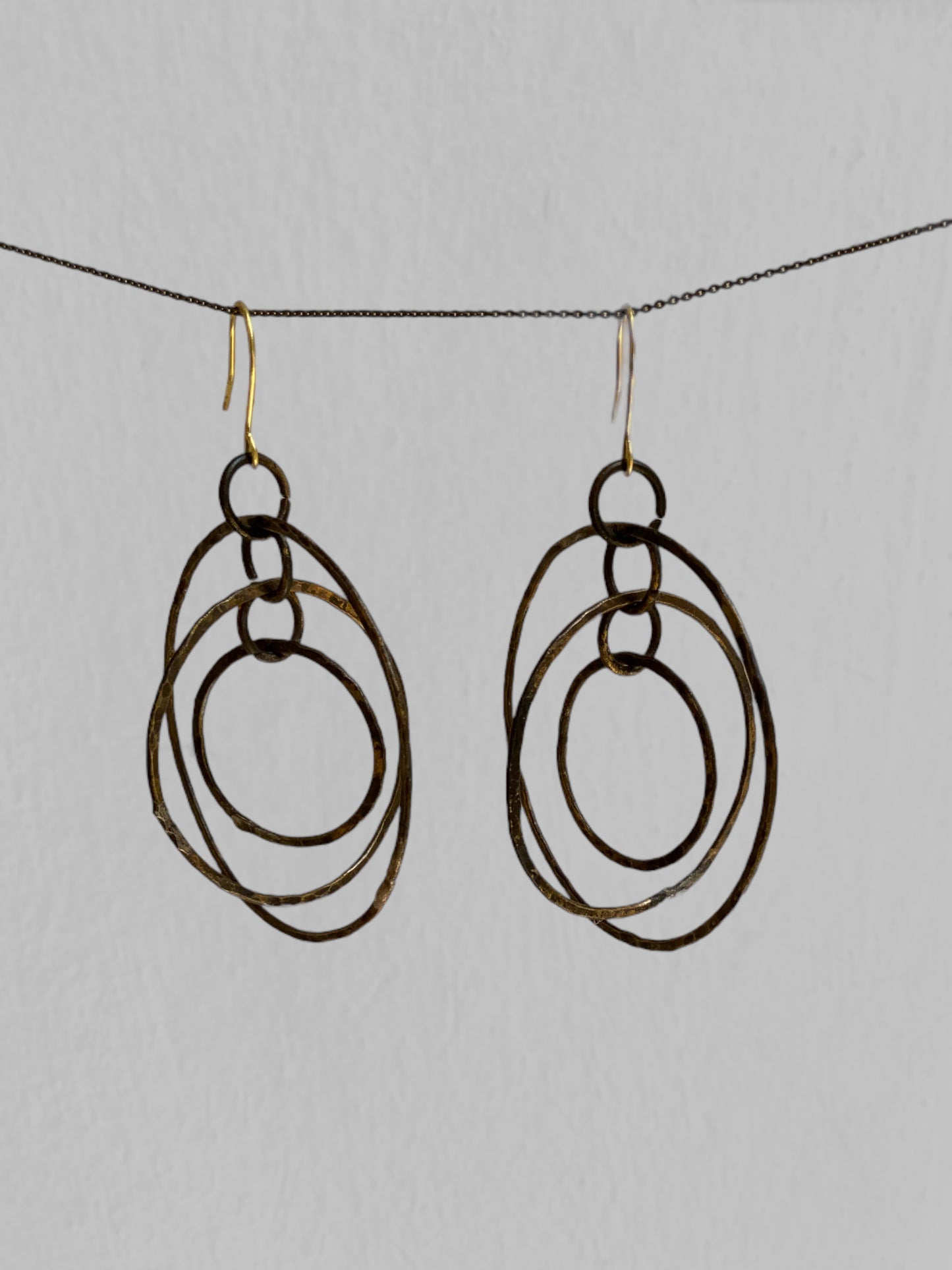 A pair of handcrafted overlapping oxidized brass hoop earrings with a hammered texture, suspended on gold hooks, styled against a neutral background for a bold and artistic design.
