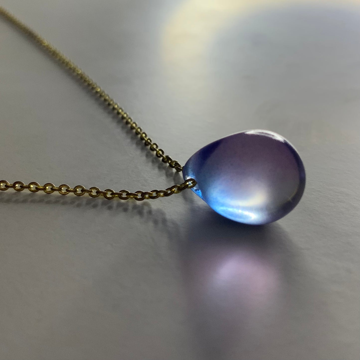 A handcrafted necklace featuring a purple gradient translucent teardrop glass pendant, suspended on a delicate brass chain, styled on a soft-lit background for a modern and elegant look.