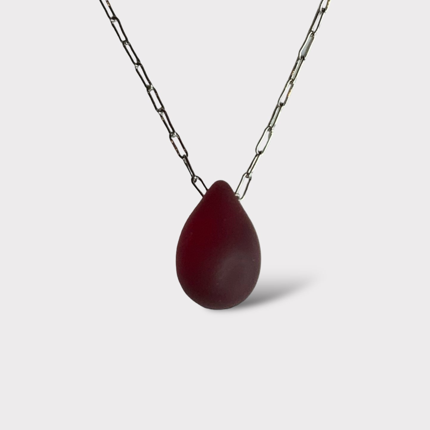 A deep ruby matte teardrop-shaped glass pendant, suspended on a stainless steel chain, styled on a clean white background for a bold and elegant look.