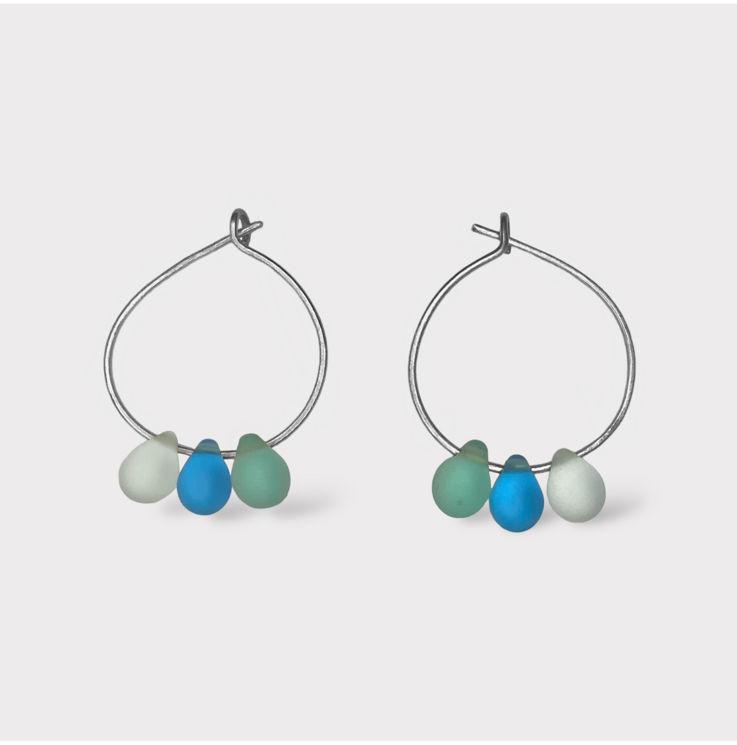 Playful silver hoop earrings adorned with aqua, turquoise, and seafoam green seaglass charms. A breezy and refreshing design reminiscent of seaside popsicles.
