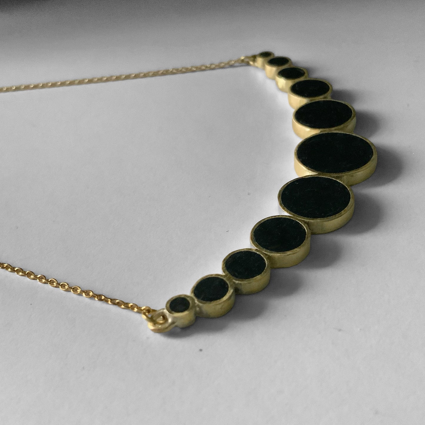 A minimalist brass necklace with black circular accents in varying sizes, arranged in a semi-circle gradient, photographed from an angle and styled on a white background for a clean look