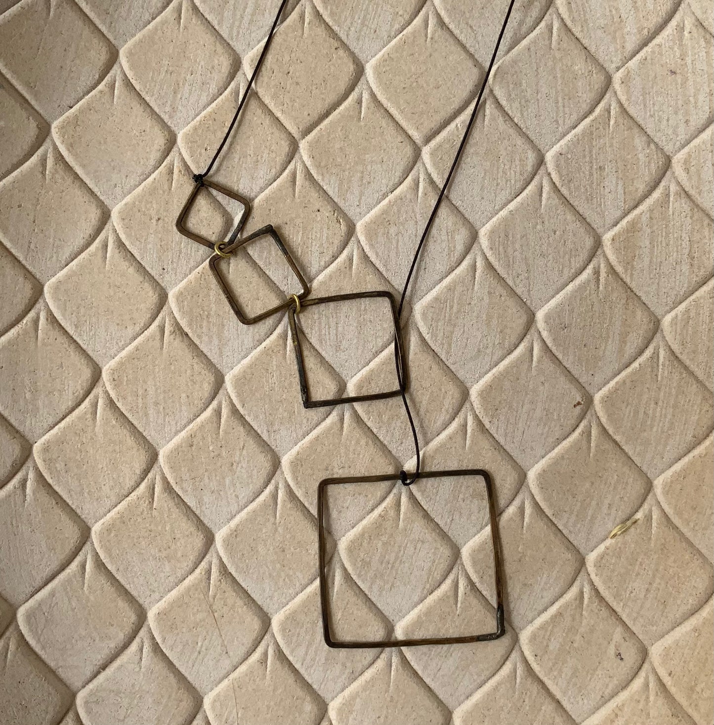 A brass lariat with cascading open square accents, styled on a beige textured background to emphasize its modern and artistic aesthetic.