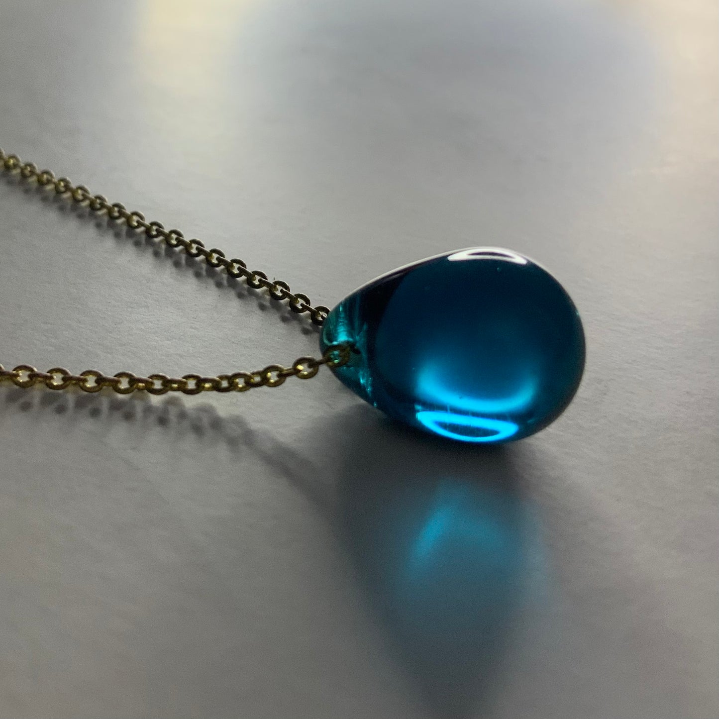 A vibrant teal translucent teardrop glass pendant, elegantly hanging from a brass chain, displayed on a softly lit surface for a fresh and minimalist design.