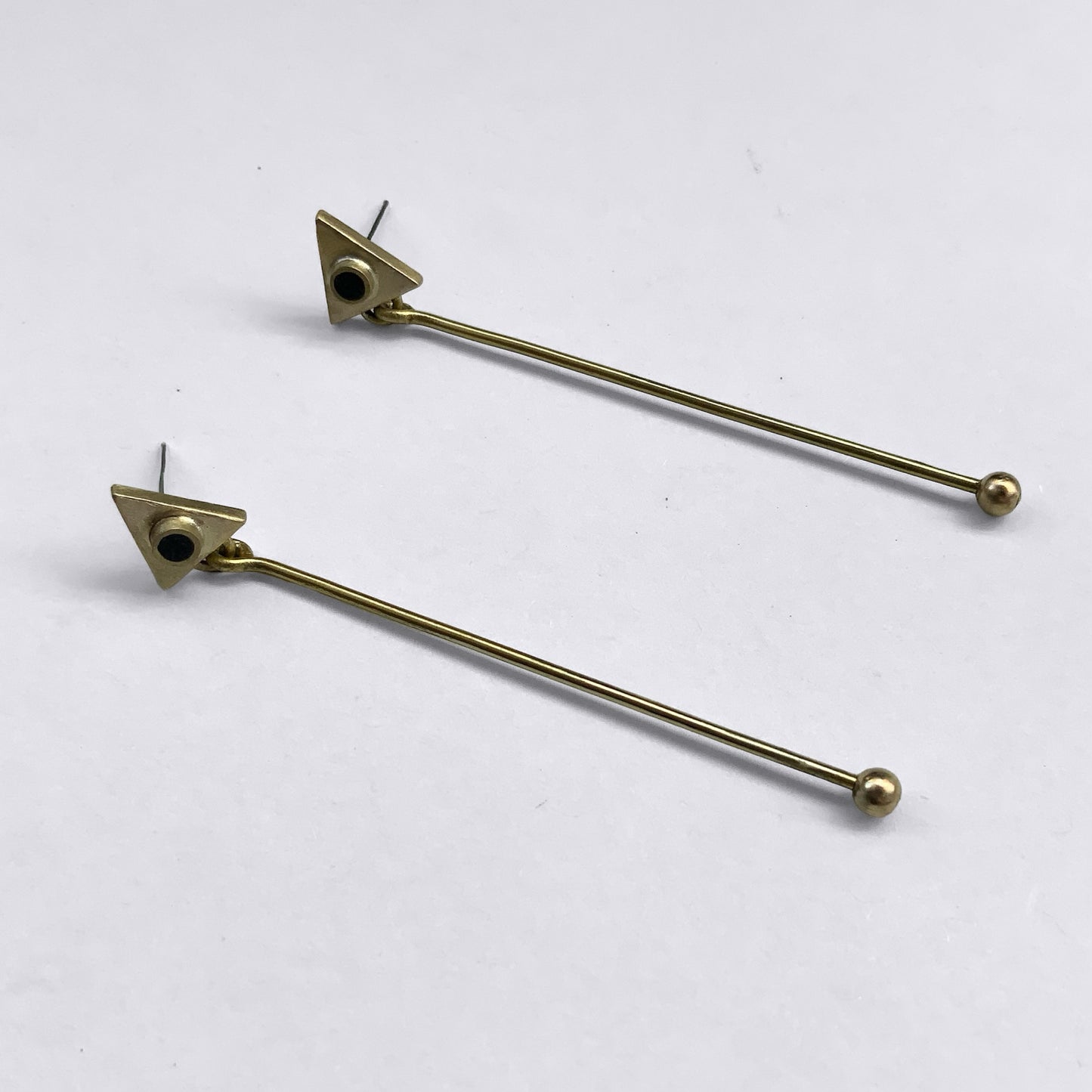 Minimalist brass earrings featuring a geometric triangle stud with a black circular accent, paired with a long, sleek brass rod ending in a small ball detail, displayed on a light background