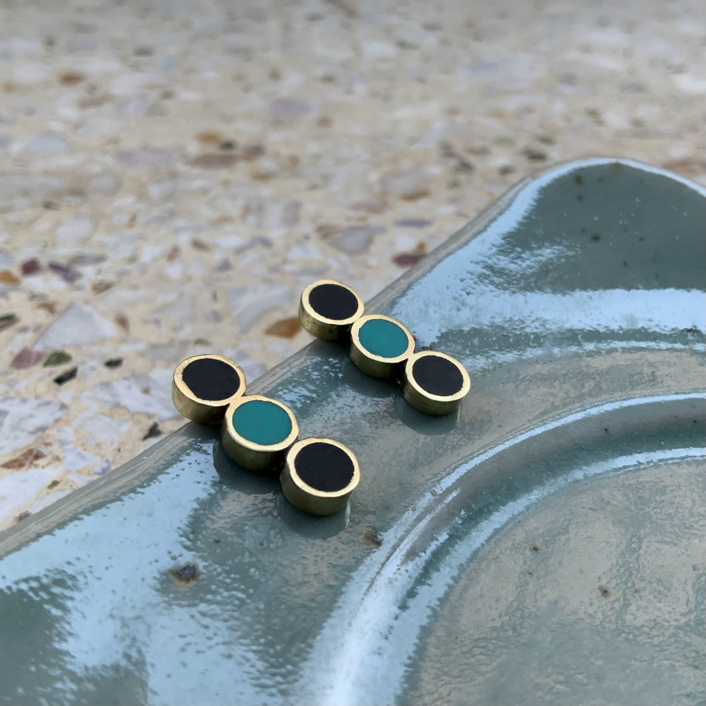A pair of brass stud earrings featuring three circular inlays, two black and a middle turquoise, arranged in a linear design, displayed on a speckled ceramic plate for an artistic presentation.
