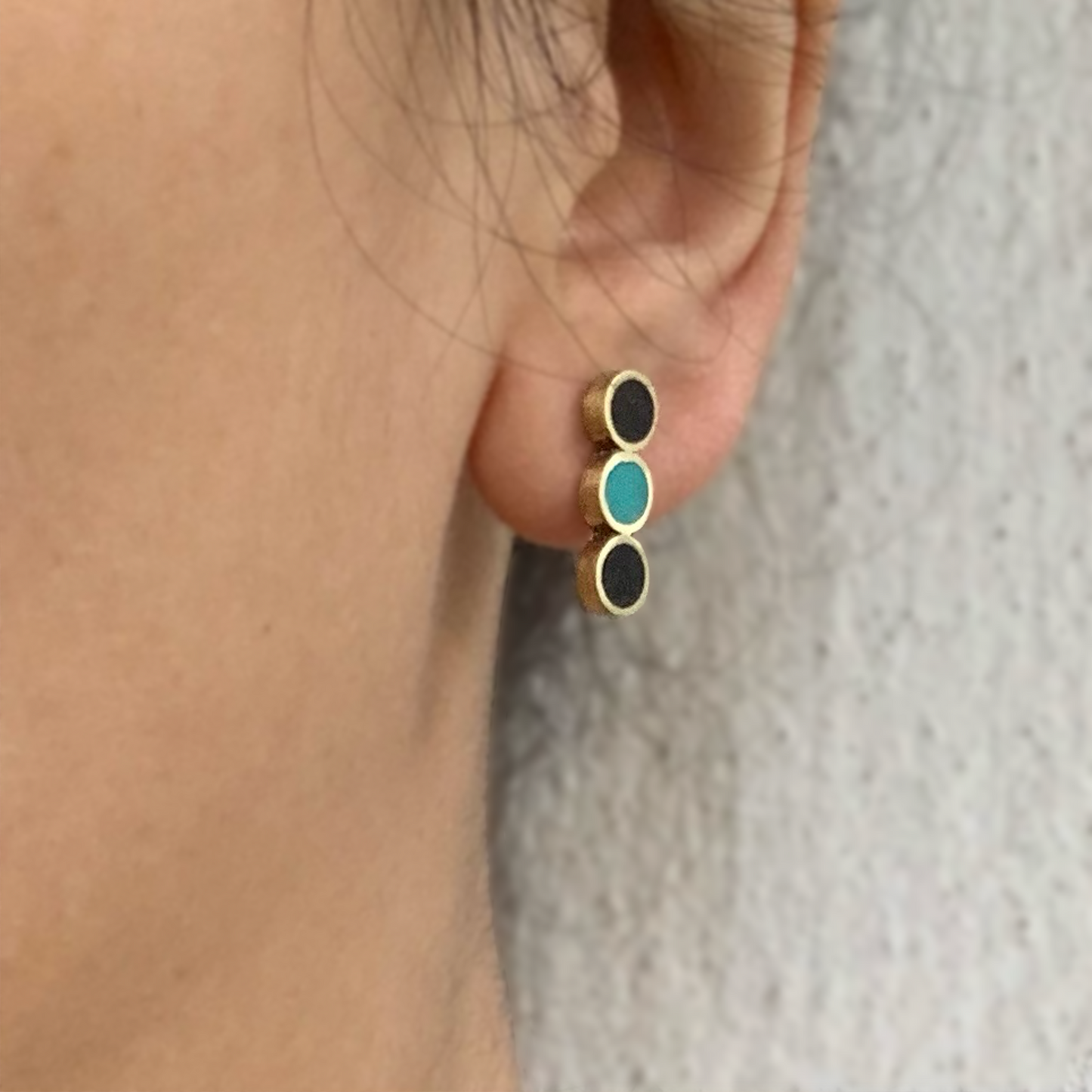 A pair of handcrafted brass stud earrings featuring three circular inlays—two black and one turquoise—worn by a model to highlight their fit and elegant design