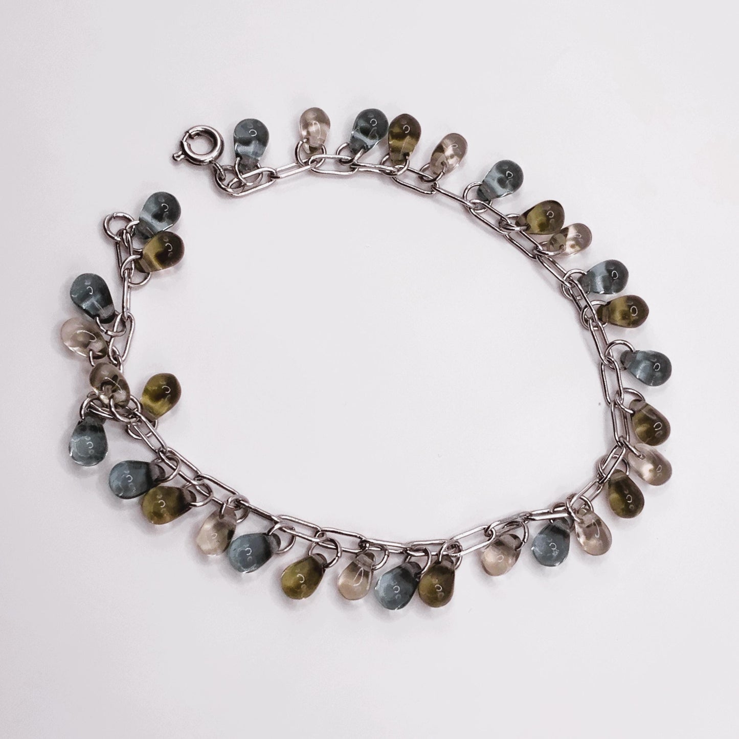 Circular silver chain bracelet with glass teardrop charms in smokey quartz, blue, and translucent, arranged flat on a white surface