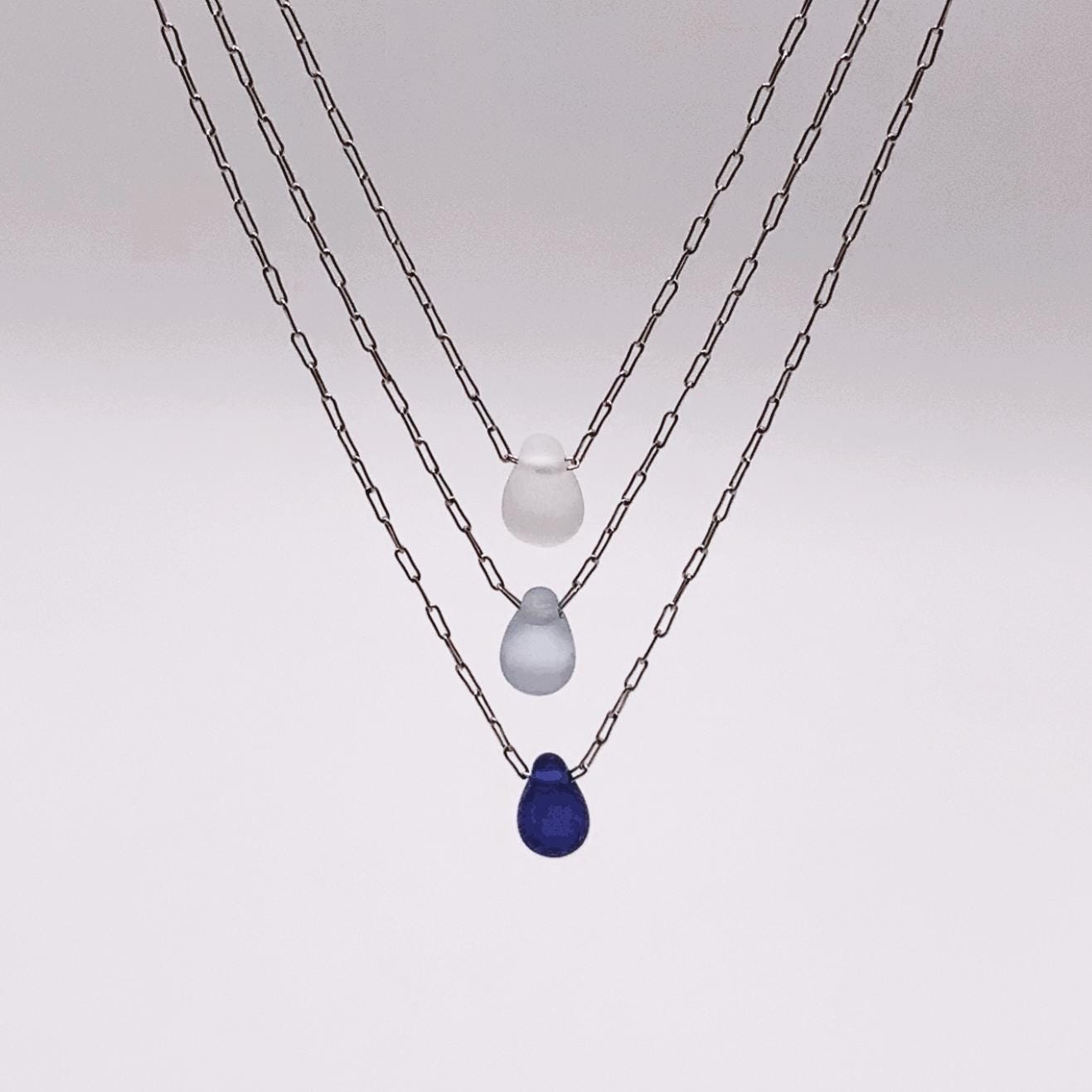 Close-up of triple-layered silver chain necklaces featuring frosted glass teardrop pendants in white, light blue, and deep blue. Summer and beach necklaces.