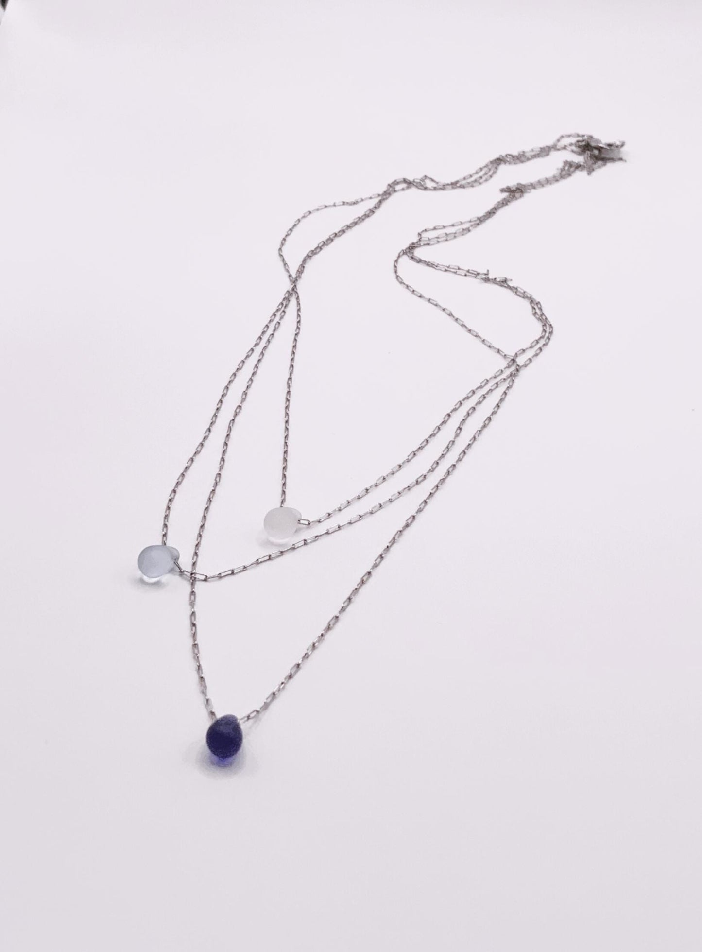 Flat-lay of three silver chain necklaces featuring frosted glass teardrop pendants in white, light blue, and deep blue on a clean white surface.