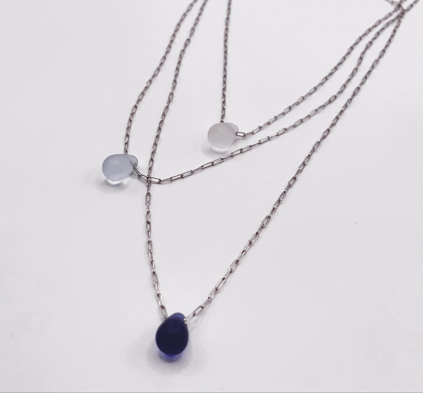 Flat-lay of triple-layered silver chain necklaces with frosted glass teardrop pendants in white, light blue, and deep blue. A sleek and versatile accessory for any occasion.