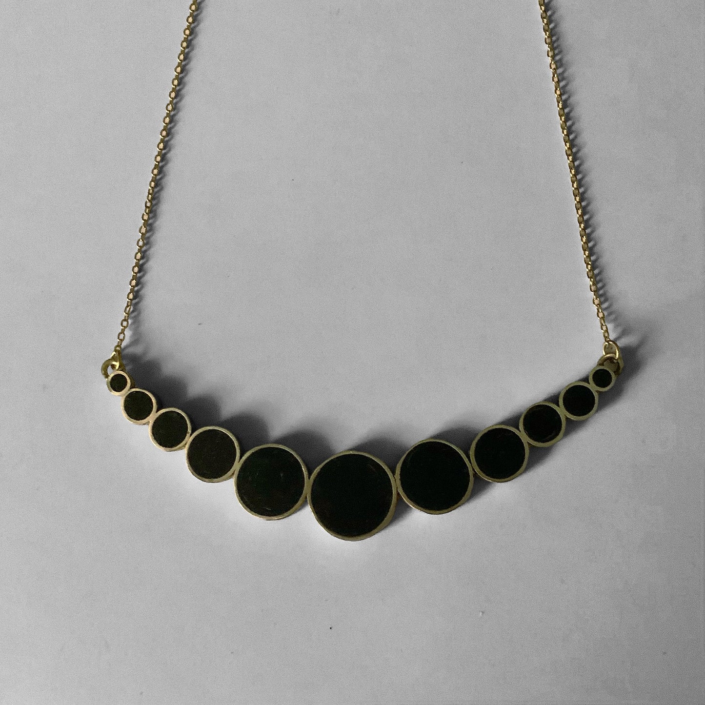 A top veiw, flatlay pictute of a brass necklace with black circular accents in a cascading gradient design, styled on a light gray background to highlight its elegant simplicity