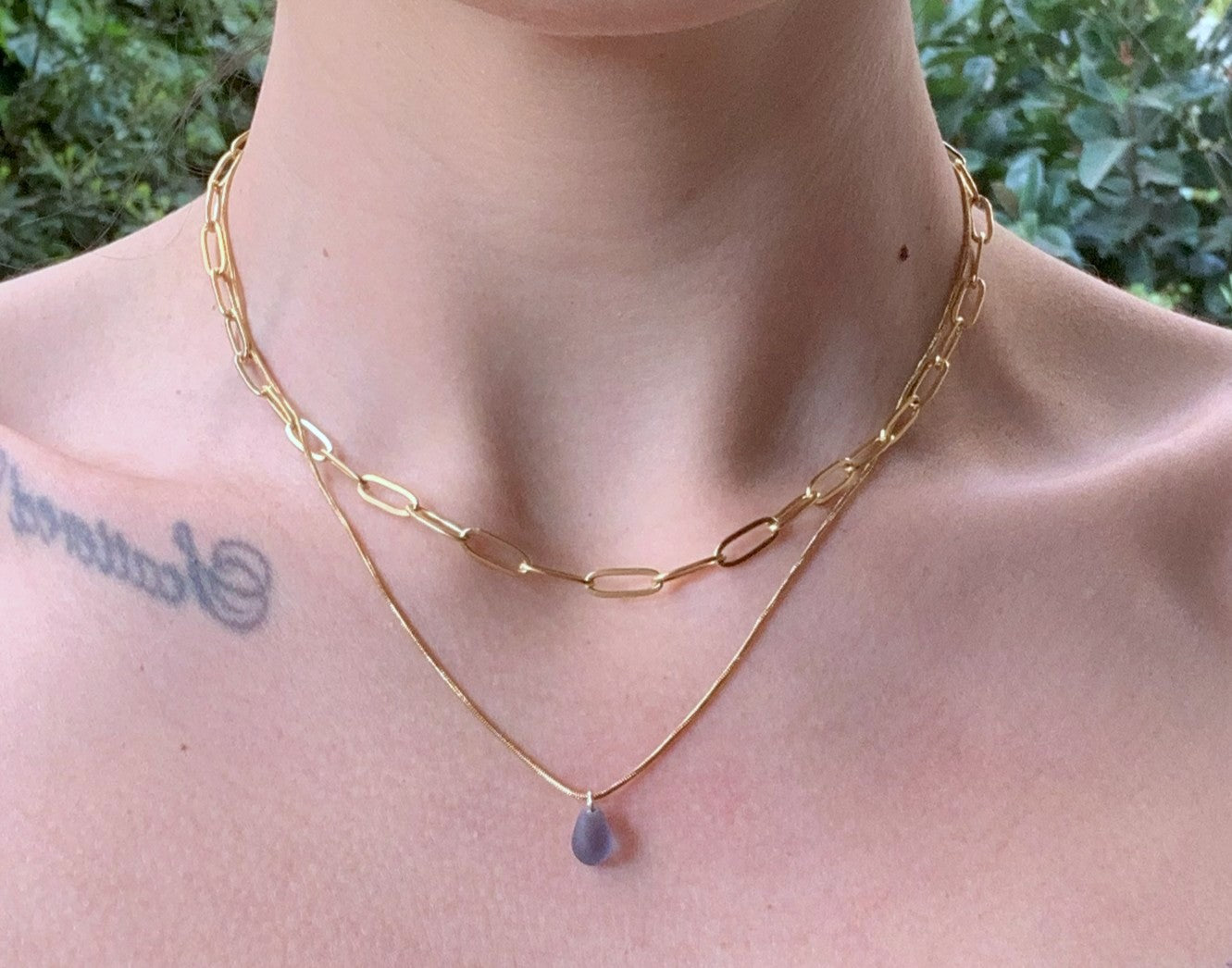Layered gold chain necklace with a lavender frosted glass teardrop pendant, featuring a chunky cable chain and a sleek snake chain. Shown on a model's neckline.
