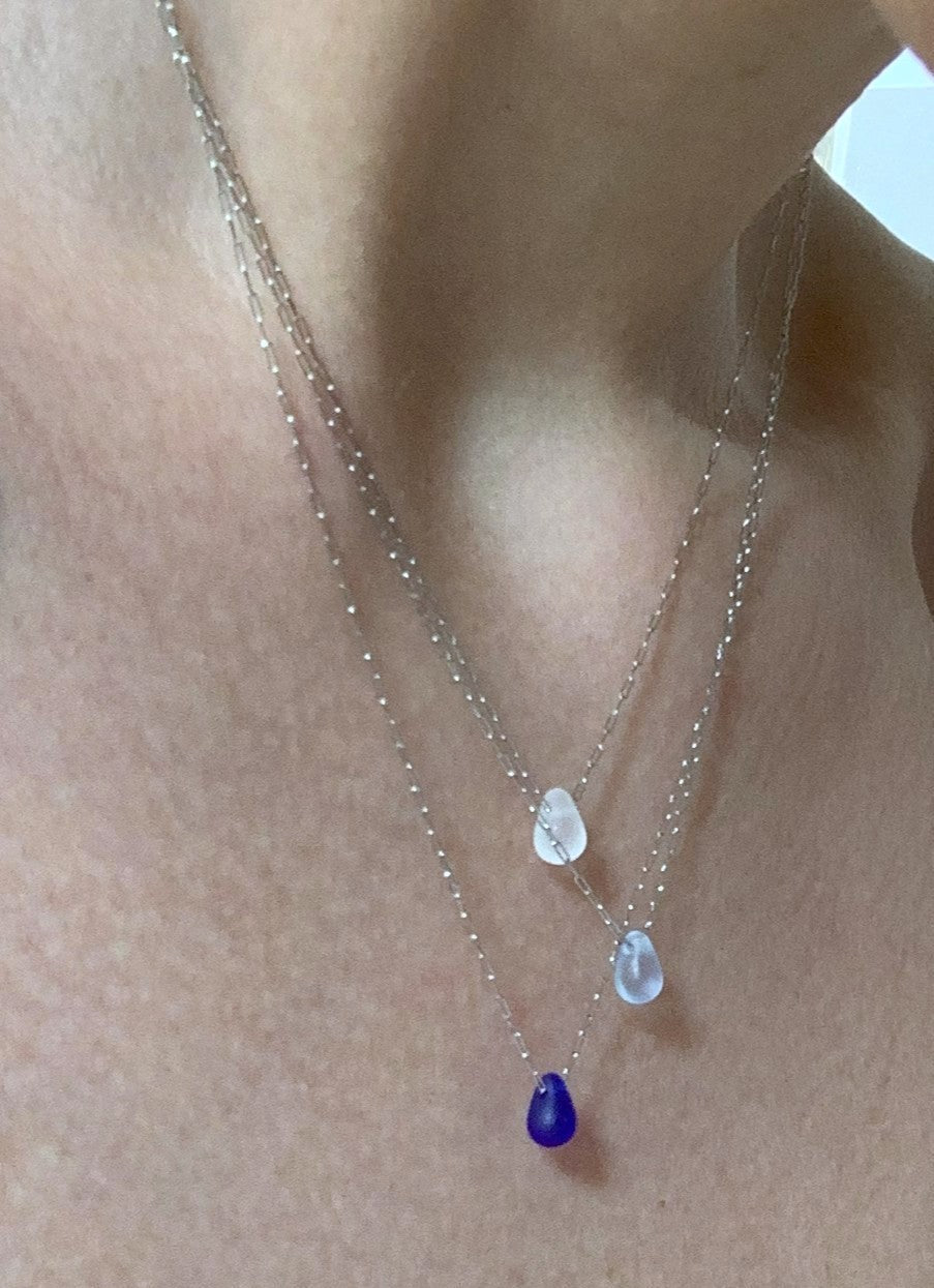 Triple layer silver chain necklace with frosted glass teardrop pendants in white, light blue, and deep blue, shown layered on a model's neckline.