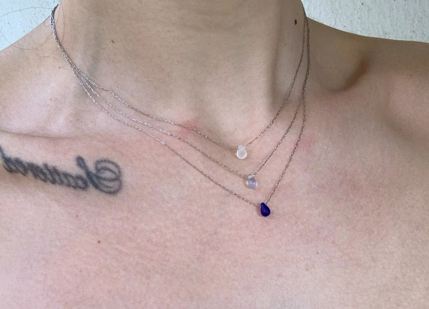 Triple-layered silver chain necklaces with frosted glass teardrop pendants in white, light blue, and deep blue, shown on a model's neckline. A delicate accessory for an elegant look.
