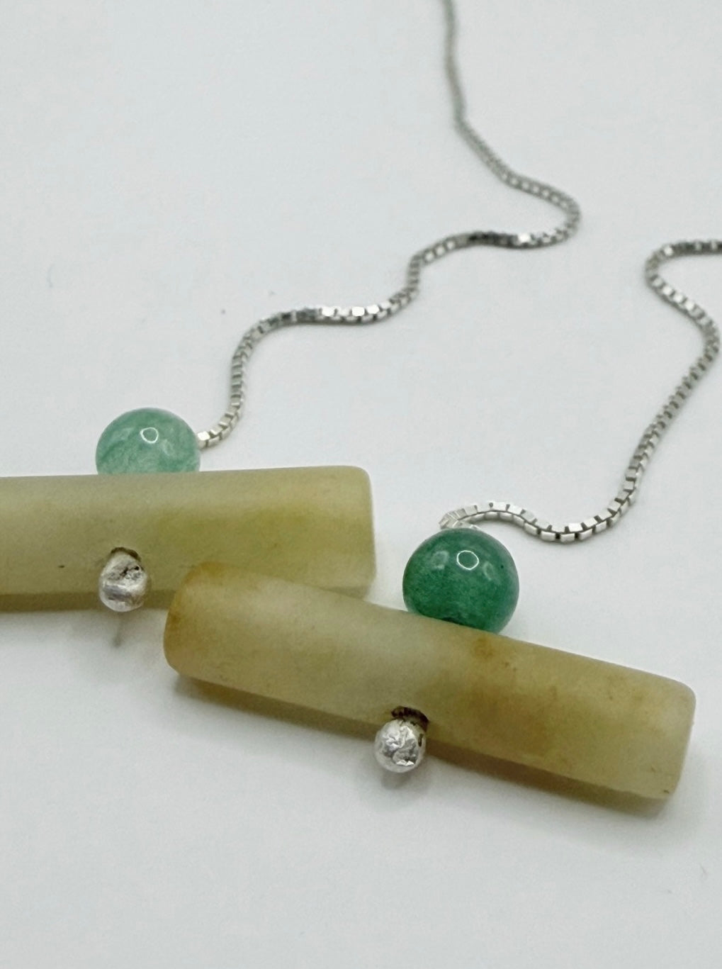 Close-up of minimalist sterling silver threader earrings with jadeite cylinder bead and aventurine green bead on a white background