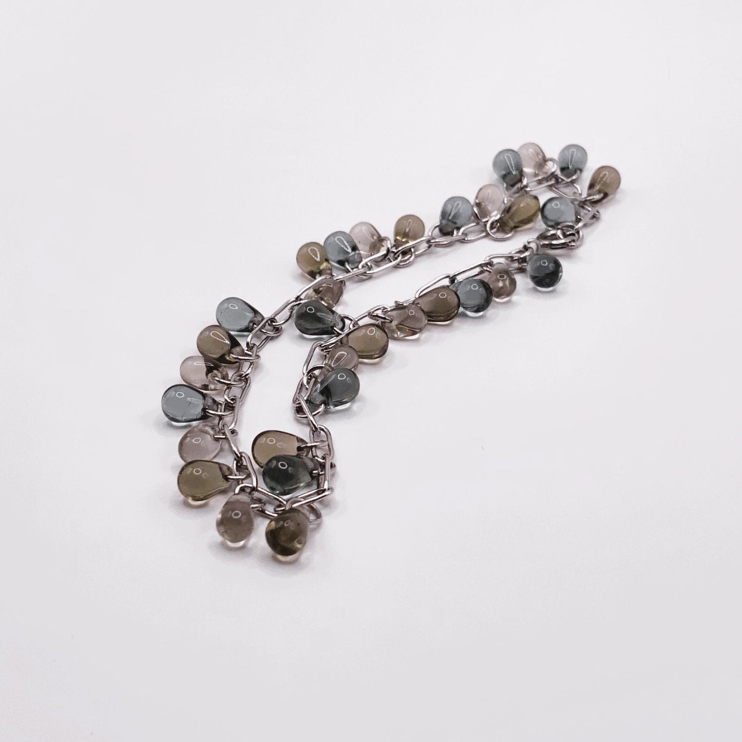 Silver chain bracelet with glass teardrop charms in earthy, blue and translucent hues, shown in a coiled arrangement on a white background.