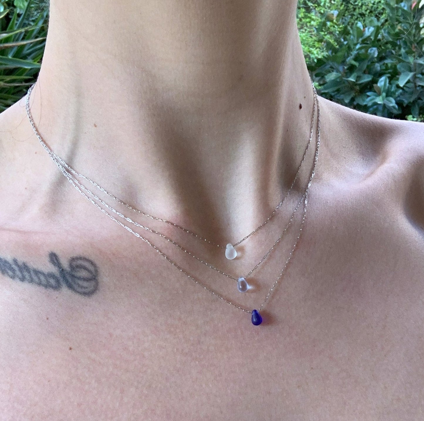 Three silver chain necklaces with frosted glass teardrop pendants in white, light blue, and deep blue, layered on a model's neckline against a natural outdoor background.