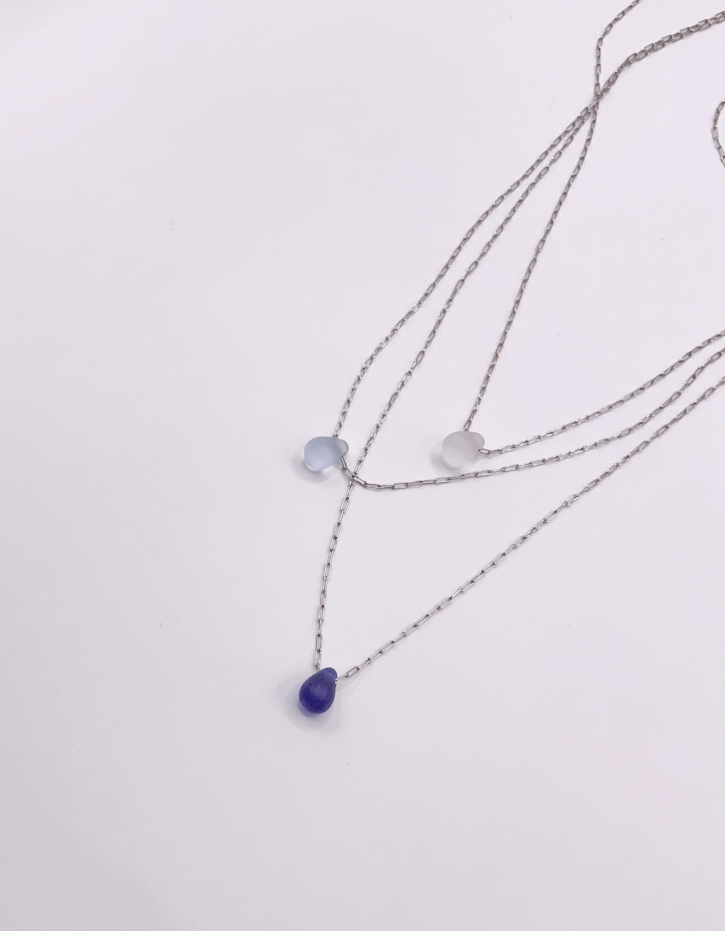 Triple silver chain necklace with frosted glass teardrop pendants in white, light blue, and deep blue, displayed flat on a white background.
