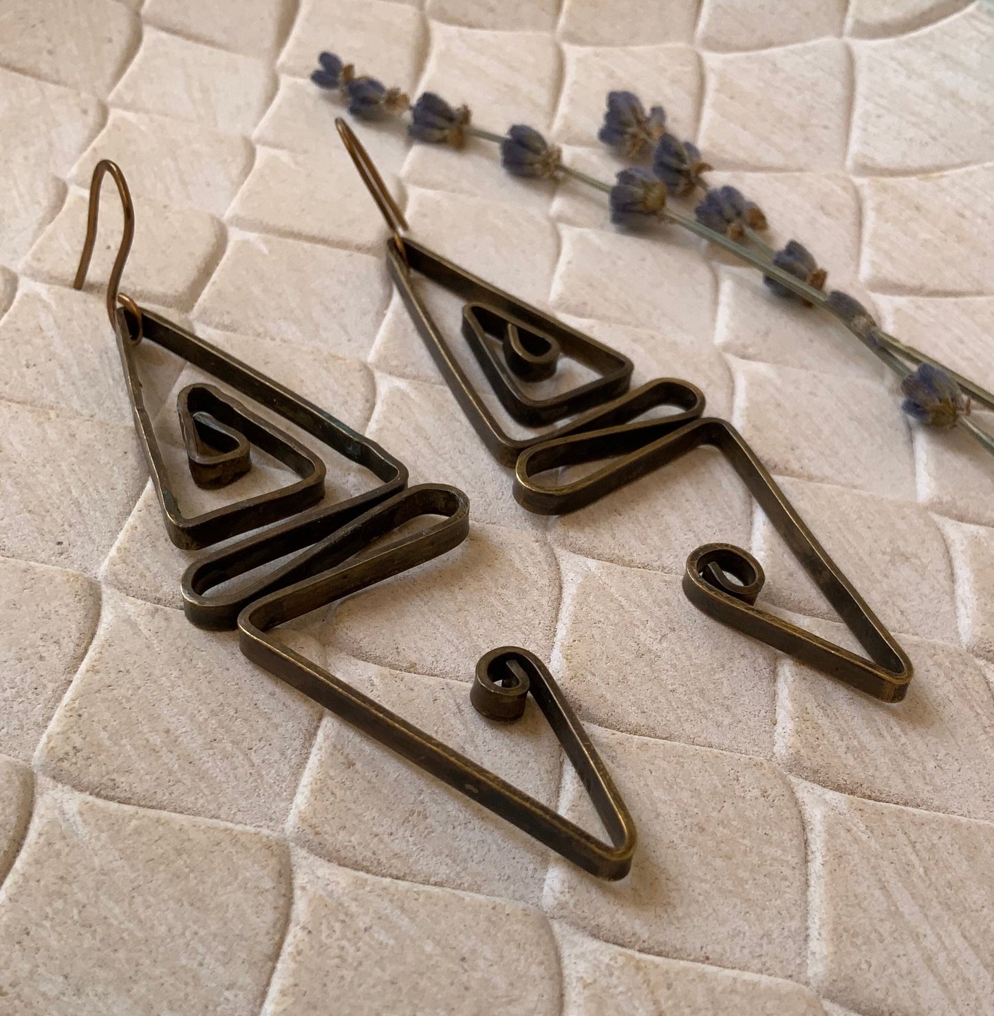 A pair of handcrafted brass geometric earrings featuring angular and spiral designs, styled on a beige textured background with sprigs of lavender for an elegant and artistic presentation.