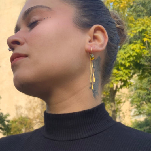 A close-up of a model wearing handcrafted brass earrings with gradient glass teardrop pendants and elongated brass bars, styled outdoors with natural lighting for an elegant and artistic presentation.