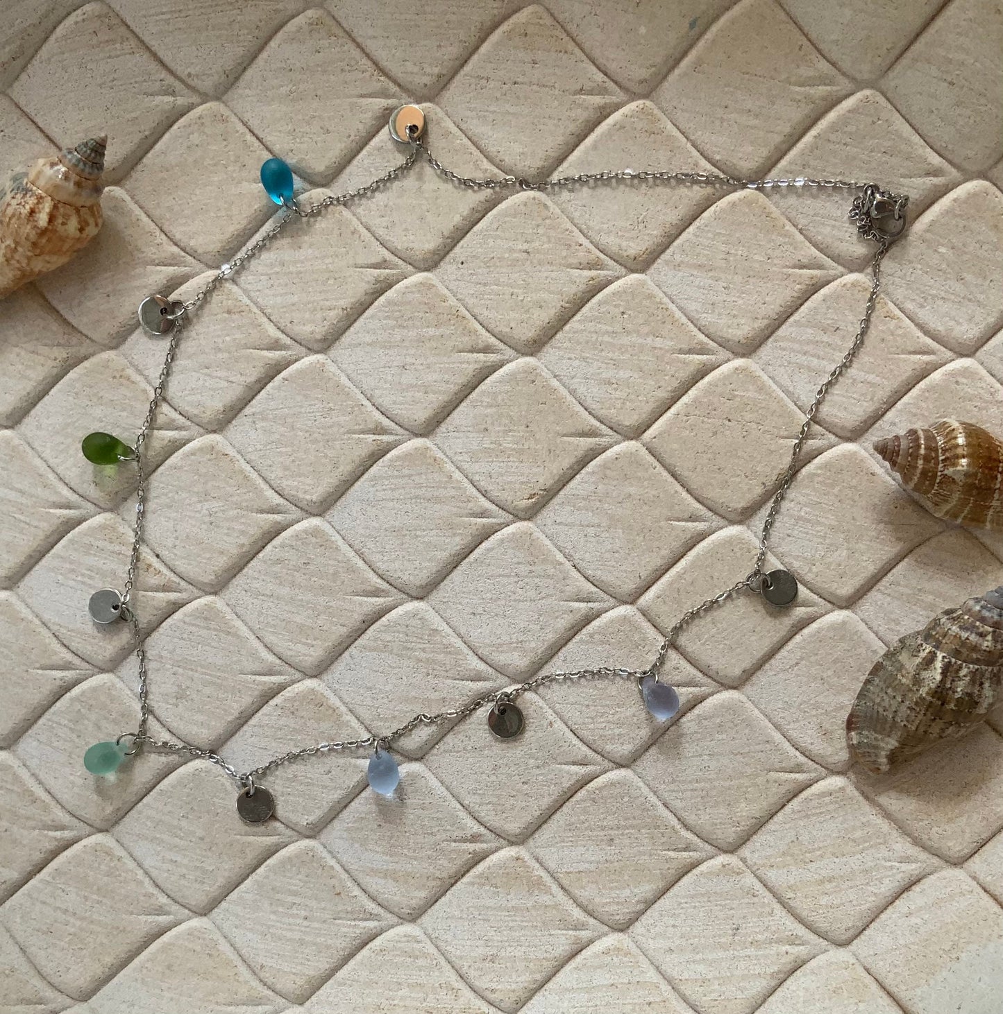 Top-view flatlay of a boho silver necklace with sea glass beads and silver coin charms on white stone tiles, handmade by Jewelry by Meow