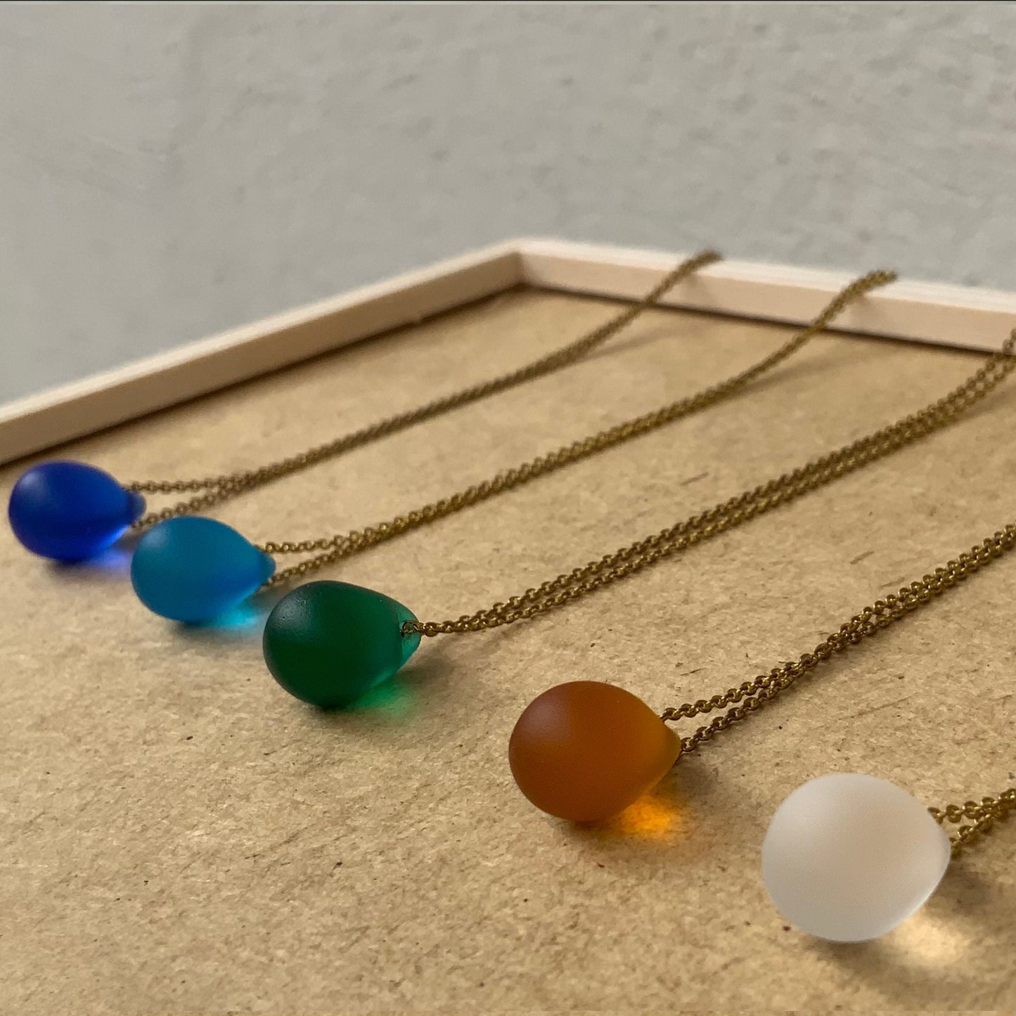 A row of matte teardrop glass pendant necklaces in bold colors, including blue, turquoise, green, amber, and frosted white, resting on a wooden surface. A minimalist and colorful collection.