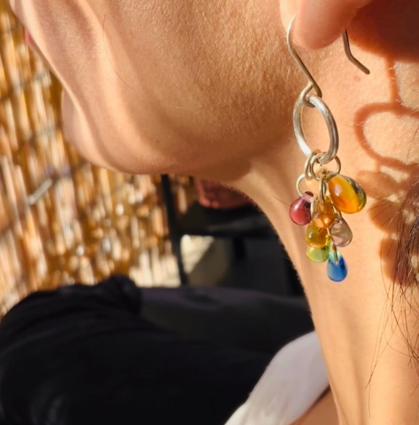 A close-up of a model wearing handcrafted silver hook earrings adorned with a vibrant cluster of multicolor glass teardrop pendants, styled in natural sunlight for a radiant and artistic presentation.