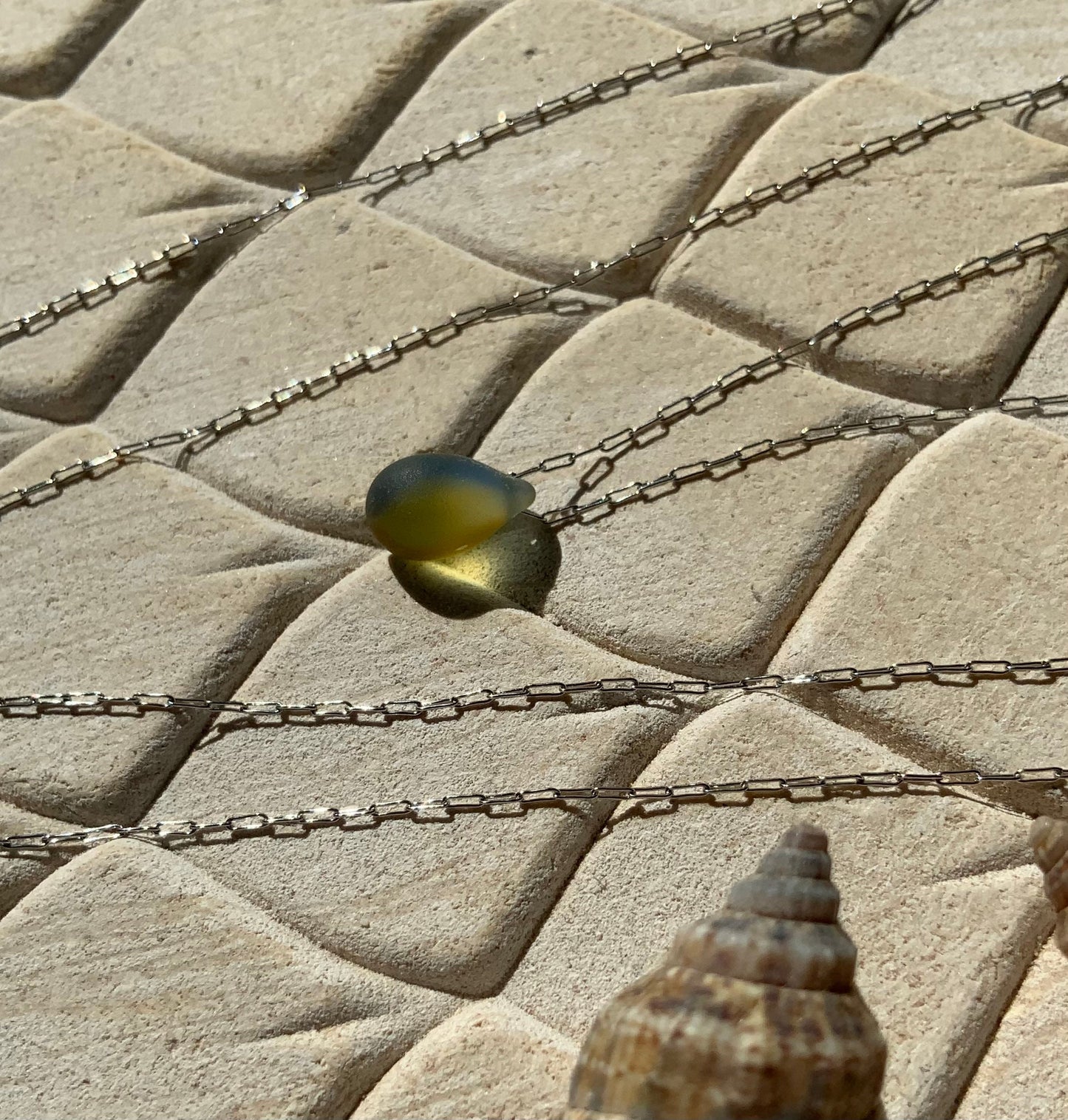 A vibrant olive green matte teardrop-shaped glass pendant, elegantly hanging from a stainless steel chain, displayed on a beige textured background with natural sunlight highlighting its bold color.