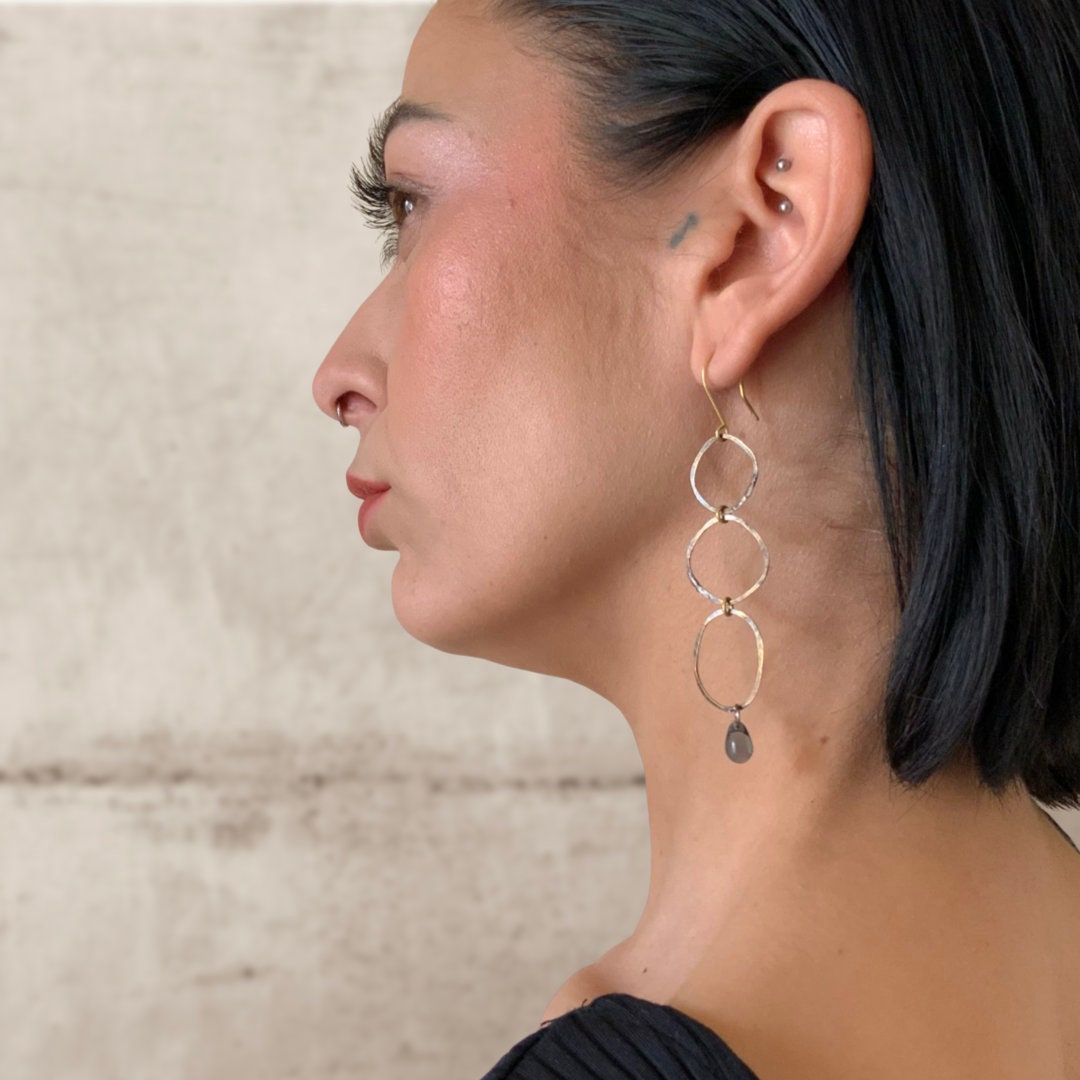A close-up of a model wearing handcrafted oval link earrings featuring interconnected hammered brass links and a blue teardrop glass pendant, styled with natural lighting for an elegant and modern look.