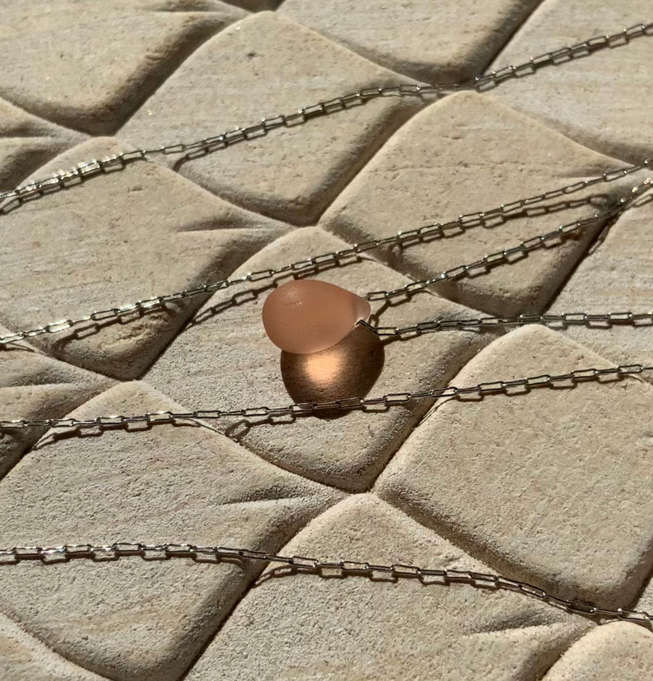 A soft peach matte teardrop-shaped glass pendant, suspended on a stainless steel chain, styled on a beige textured background with natural sunlight enhancing its warm tones.