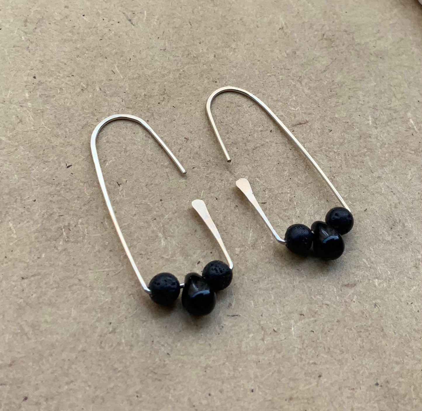 A pair of handcrafted silver hoop earrings featuring a black glass and two lava stone beads on each hoop, designed for a bold yet minimalist statement. Top view displayed on a light wooden surface creating a clean minimalist style