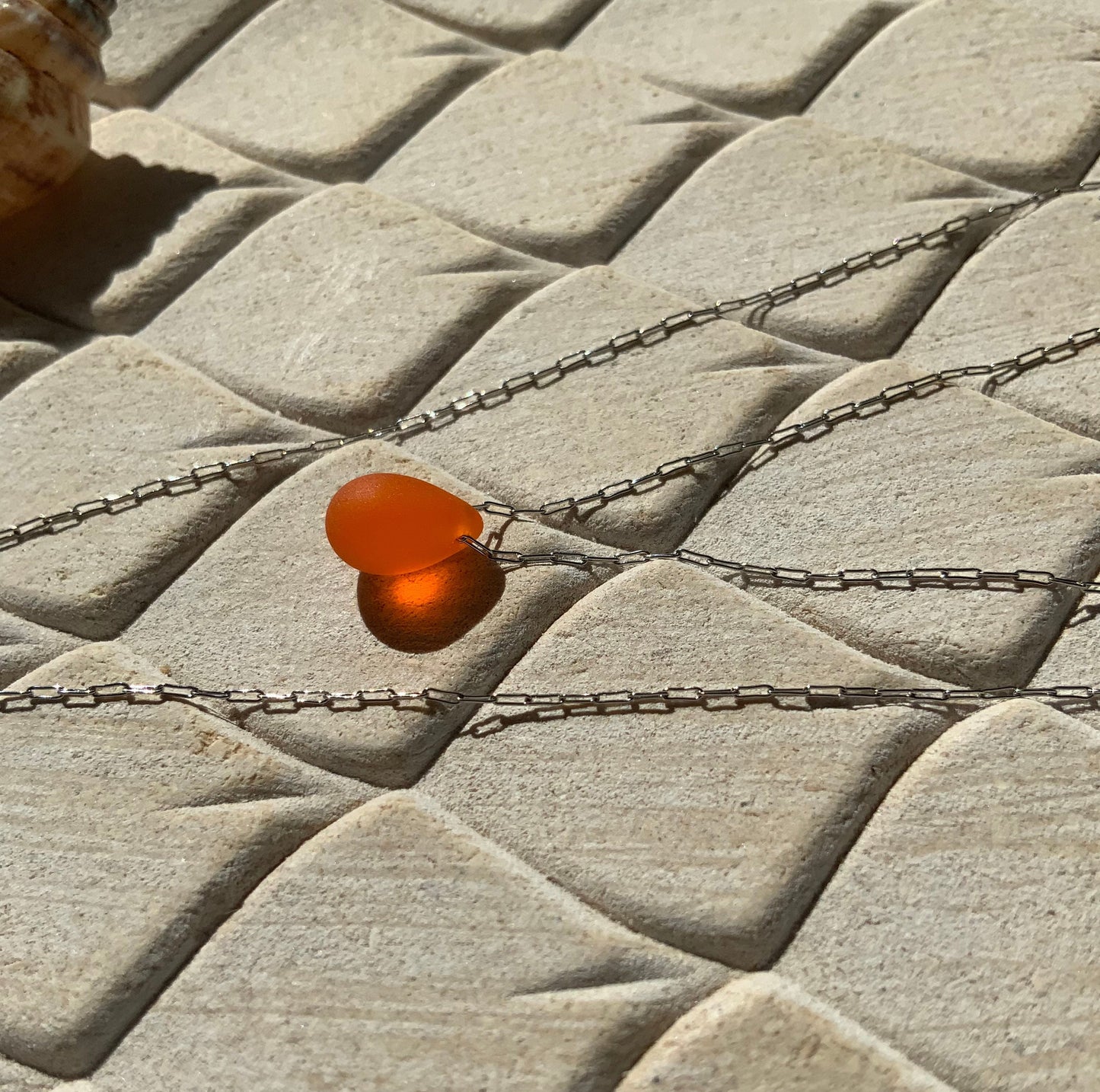 A vibrant tangerine teardrop-shaped glass pendant, styled on a stainless steel chain and displayed on a beige textured background for an artistic presentation.