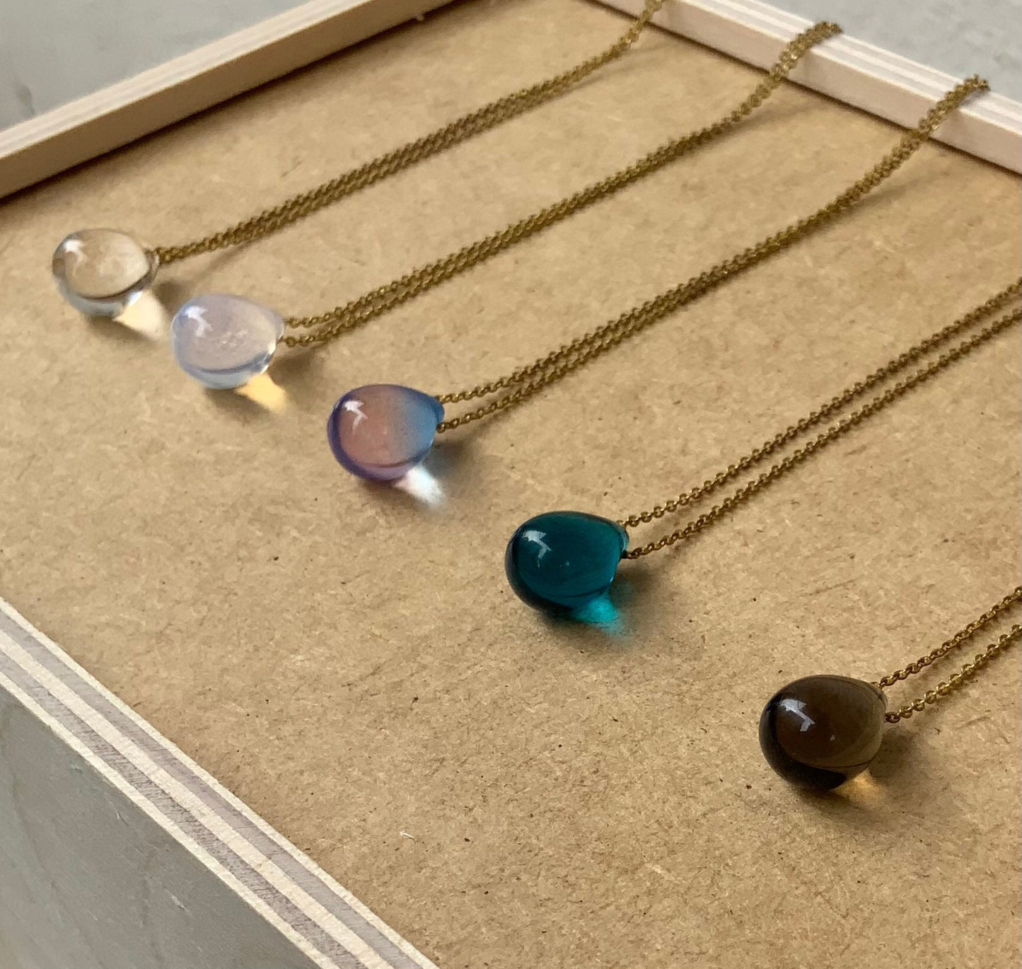 A set of teardrop glass pendant necklaces in various translucent shades, including white, purple, aqua blue, and brown, displayed on a beige wooden surface for an artistic and cohesive presentation
