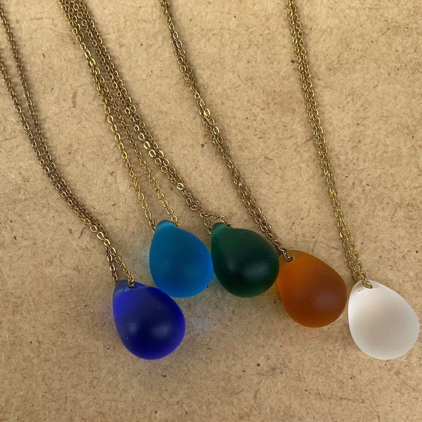 Flat lay of a collection of matte teardrop pendant necklaces in cobalt blue, turquoise, emerald green, amber and frosted white, displayed on a wooden board. Each pendant is handcrafted and suspended on a stainless steel chain for a cohesive and elegant presentation.
