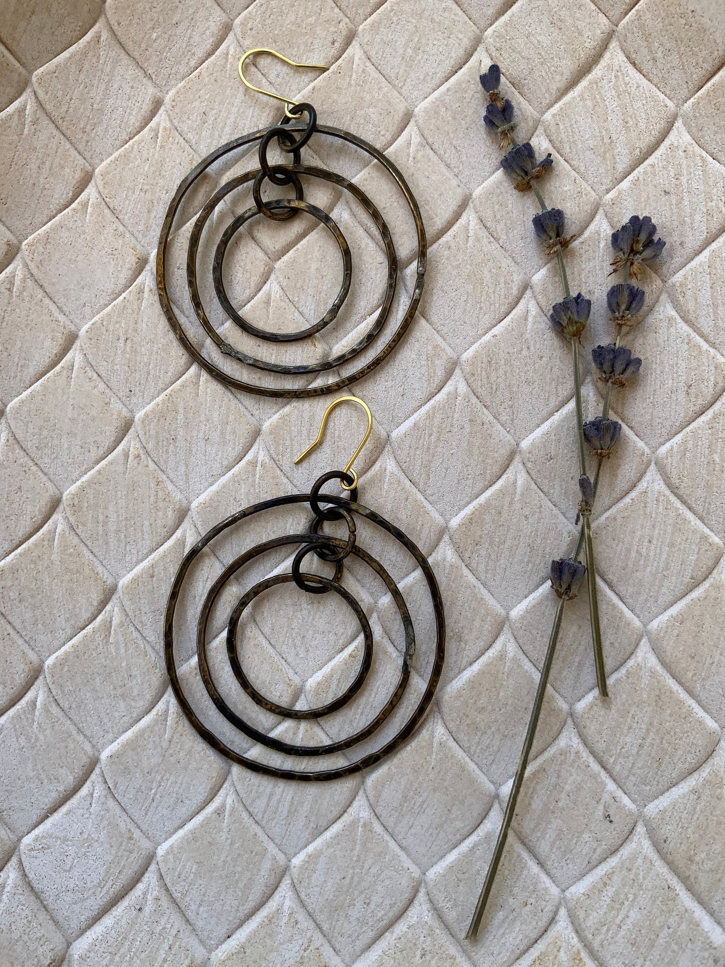 A pair of hammered brass triple circle hoop earrings, displayed on a beige textured background alongside sprigs of lavender for a rustic and elegant presentation.