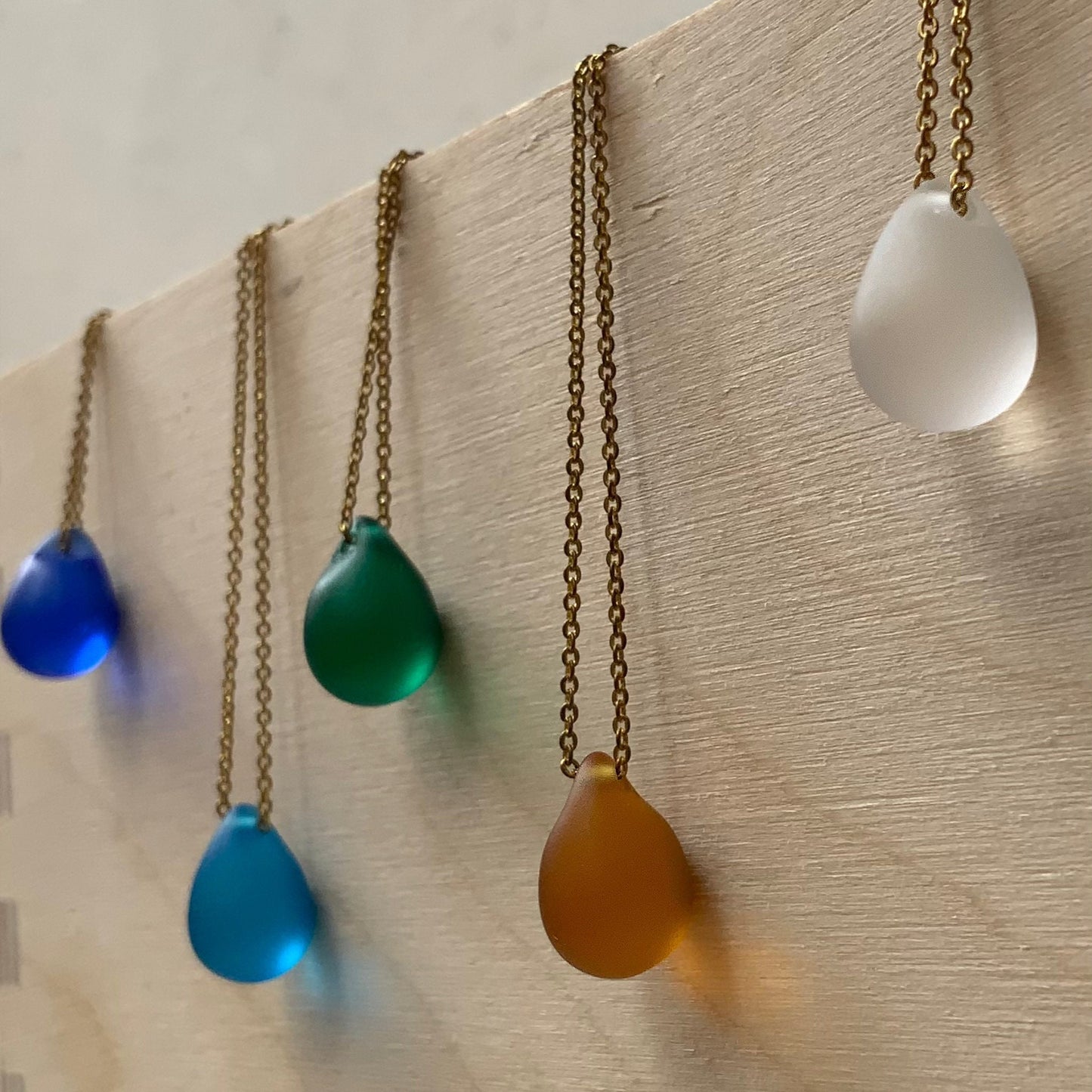 A vertical display of matte teardrop glass pendant necklaces in cobalt blue, turquoise, emerald green, amber, and frosted white, elegantly suspended on stainless steel chains against a wooden background.