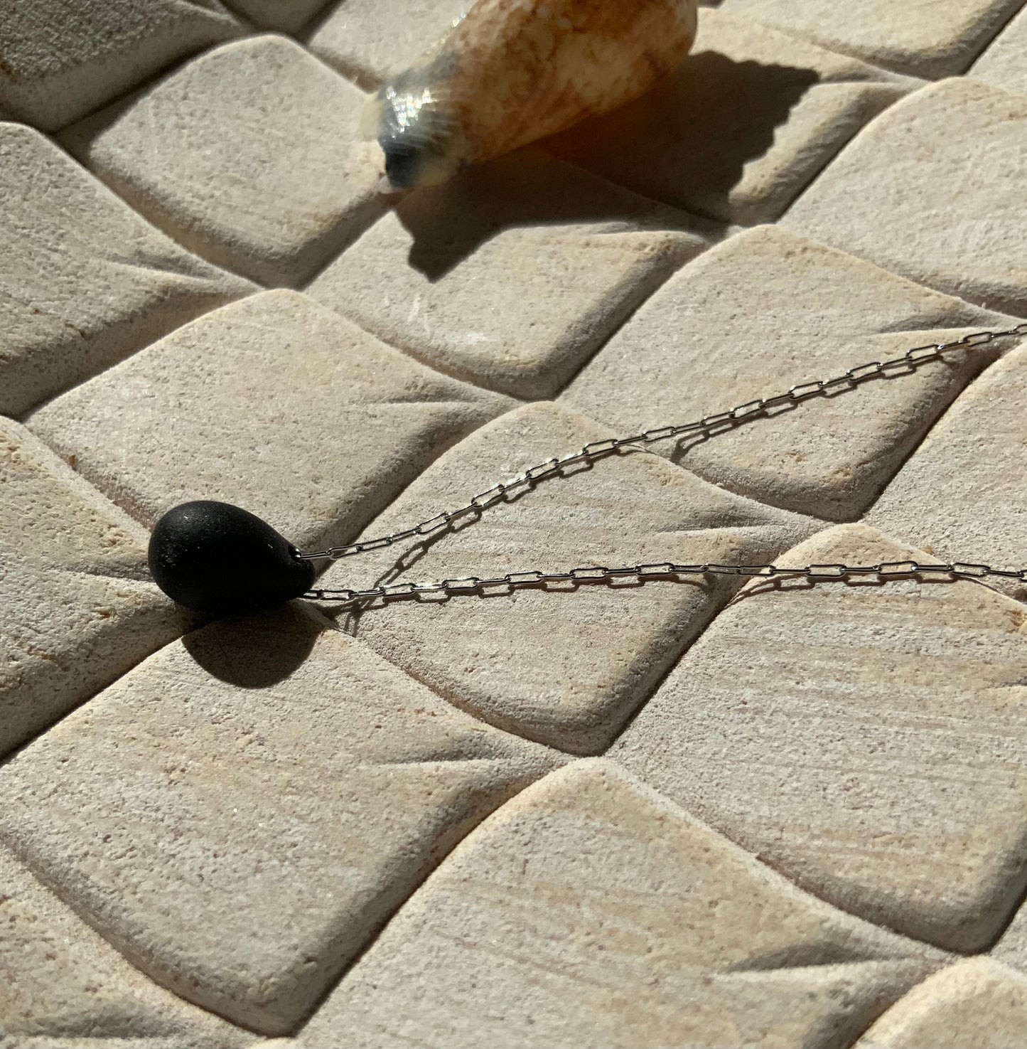 A sleek black matte teardrop-shaped glass pendant, suspended on a stainless steel chain, styled on a beige textured background with natural sunlight creating subtle shadows.