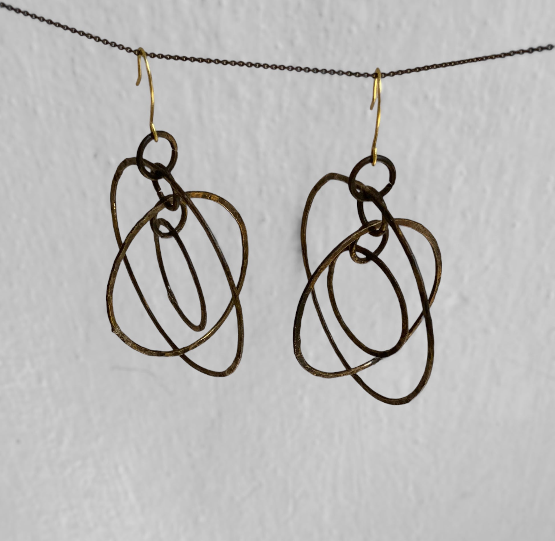 A pair of handcrafted earrings featuring abstract hammered brass loops, suspended on gold hooks, styled on a neutral background to highlight their artistic and unconventional design.