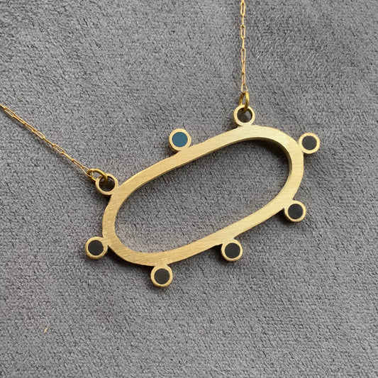 A handcrafted brass necklace featuring an elongated oval pendant with circular accents, including a single blue accent, suspended on a delicate golden stainless steel chain, displayed on a textured gray background.
