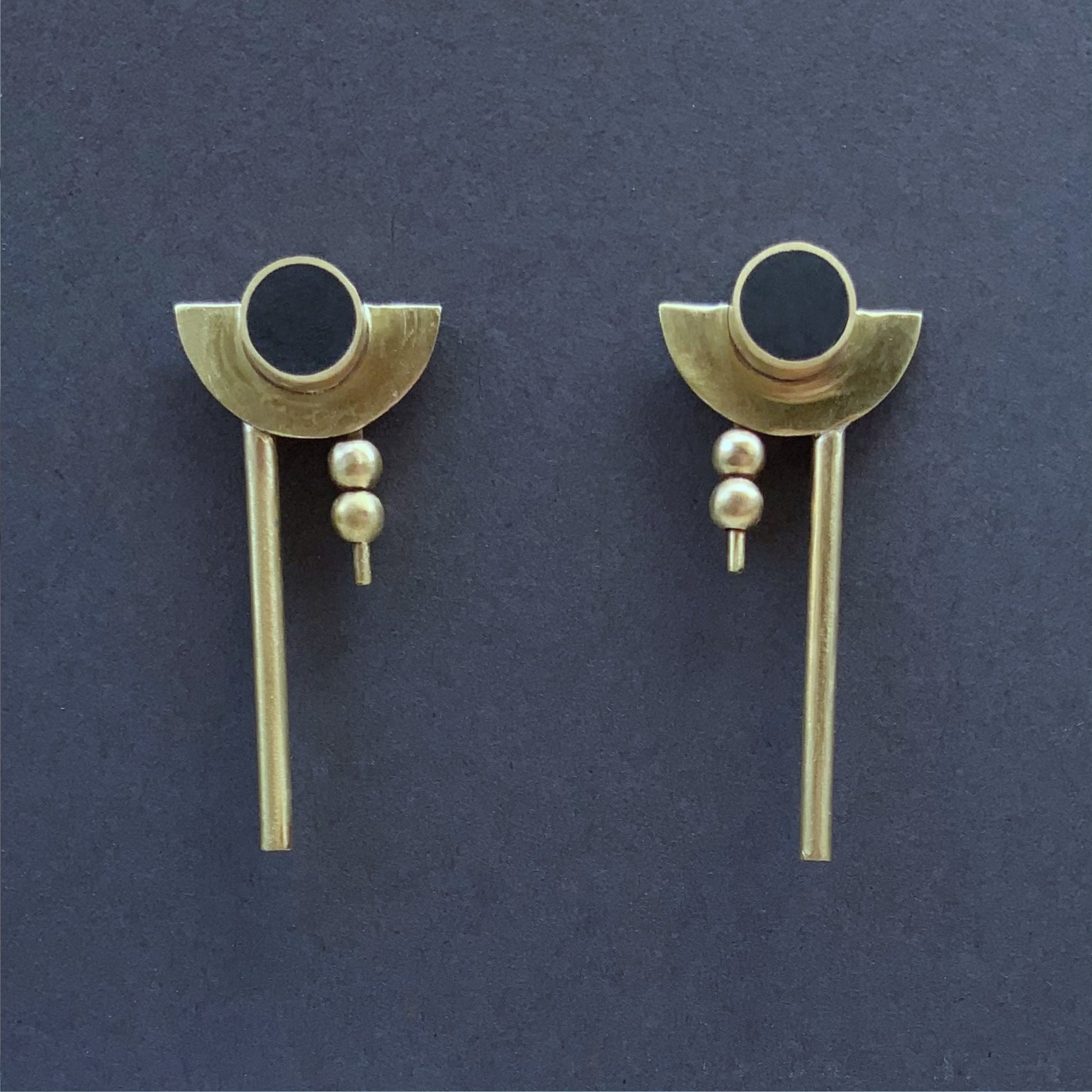 Bauhaus earrings, front view of pair of handcrafted earrings featuring a black circular centerpiece encased in brass, complemented by crescent-shaped brass accents, two small brass beads, and a sleek rod drop design, displayed on a dark background.