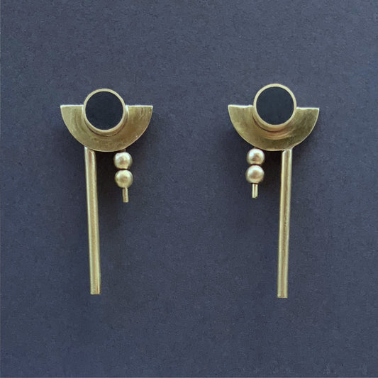 Bauhaus earrings, front view of pair of handcrafted earrings featuring a black circular centerpiece encased in brass, complemented by crescent-shaped brass accents, two small brass beads, and a sleek rod drop design, displayed on a dark background.