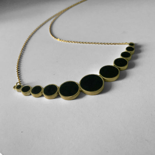 A handcrafted brass necklace featuring a gradient design of black circular accents in varying sizes, arranged in a semi-curve, suspended on a delicate staiknless steel chain, displayed on a light background.