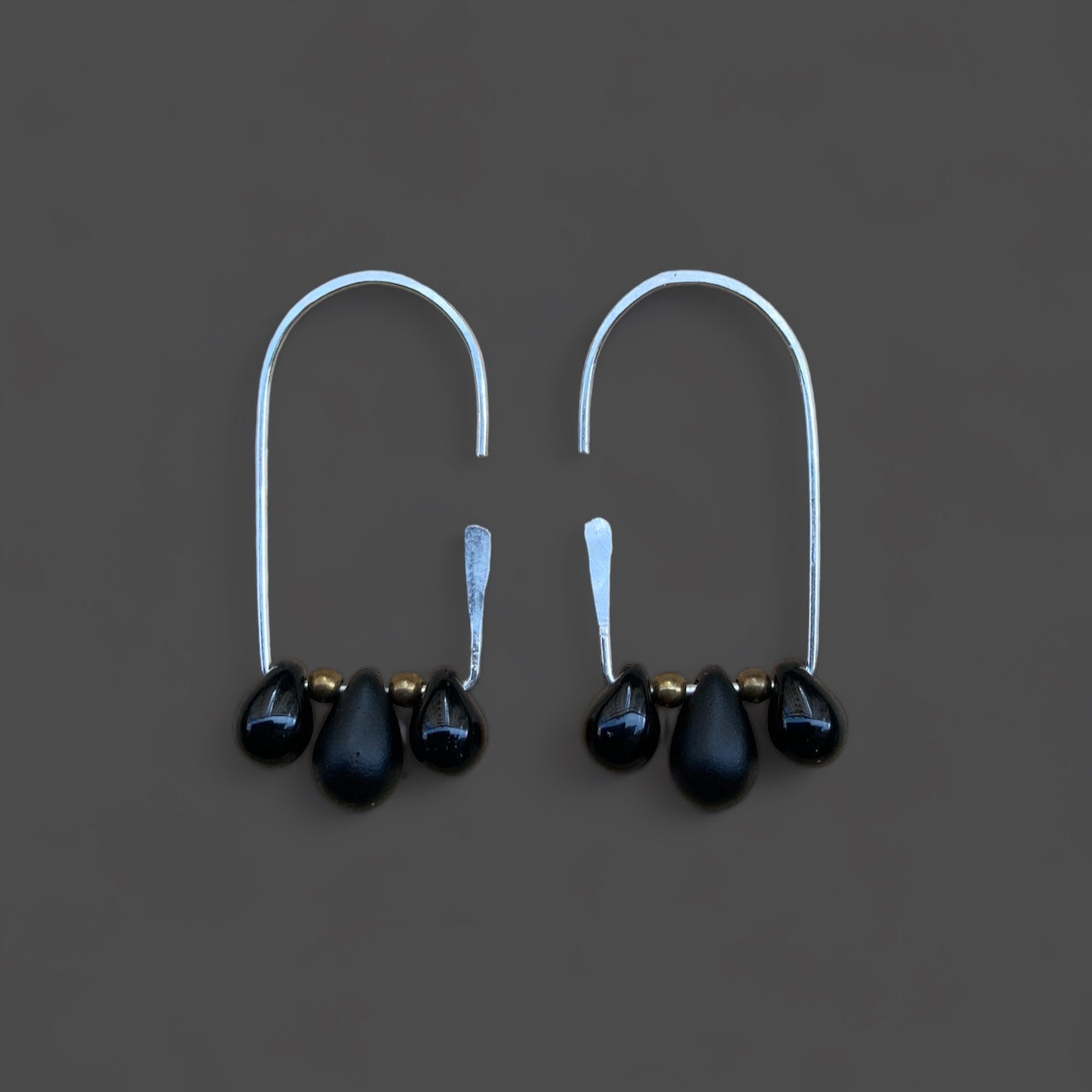 Handcrafted silver hoop earrings adorned with three black teardrop glass pendants and brass bead accents, displayed on a dark gray background. A bold and minimalist design perfect for elegant occasions.