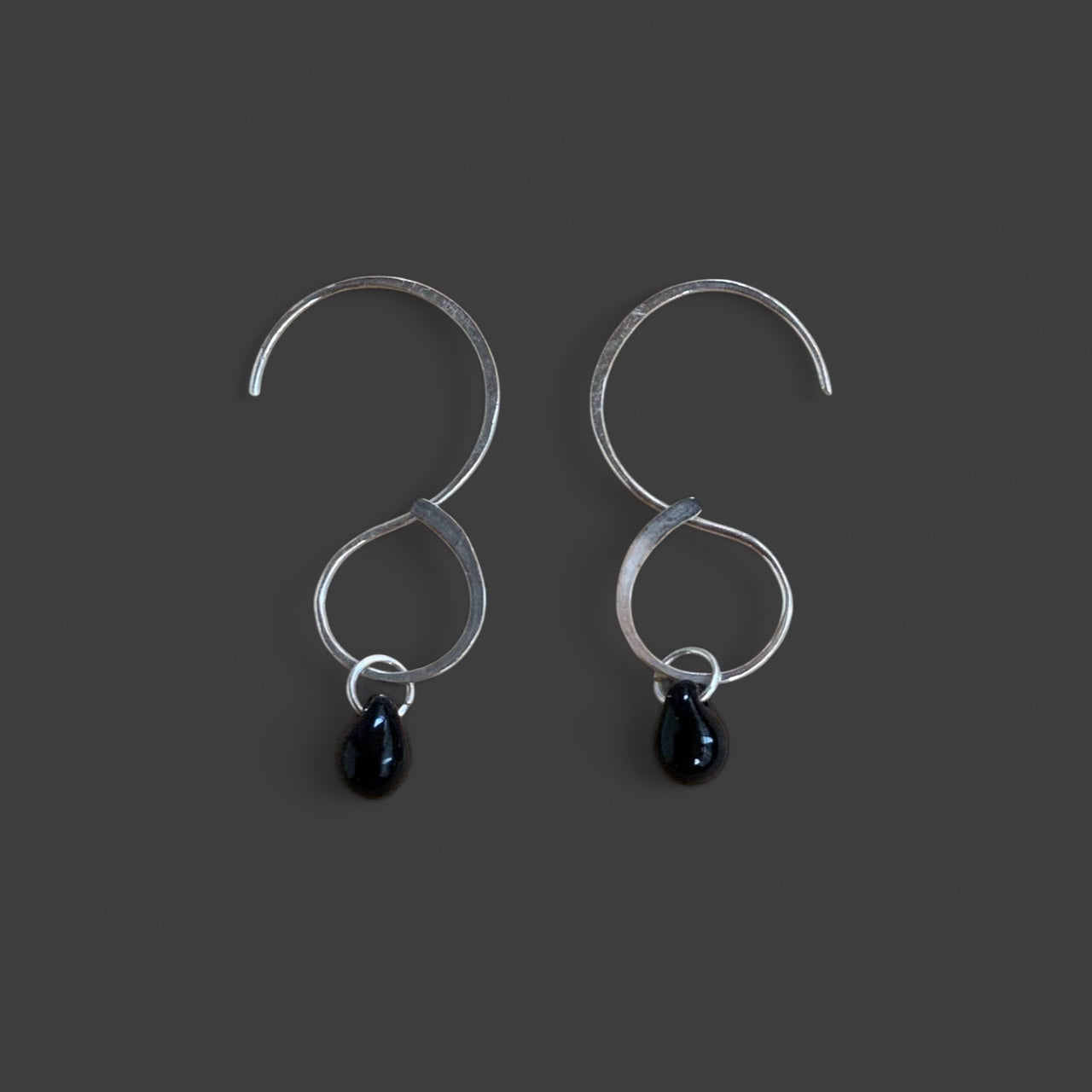 A pair of silver infinity hoop earrings with black teardrop glass pendants, displayed on a dark gray background. A minimalist and versatile accessory for a bold yet refined look.