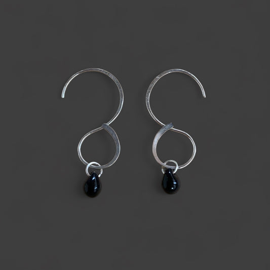 A pair of silver infinity hoop earrings with black teardrop glass pendants, displayed on a dark gray background. A minimalist and versatile accessory for a bold yet refined look.