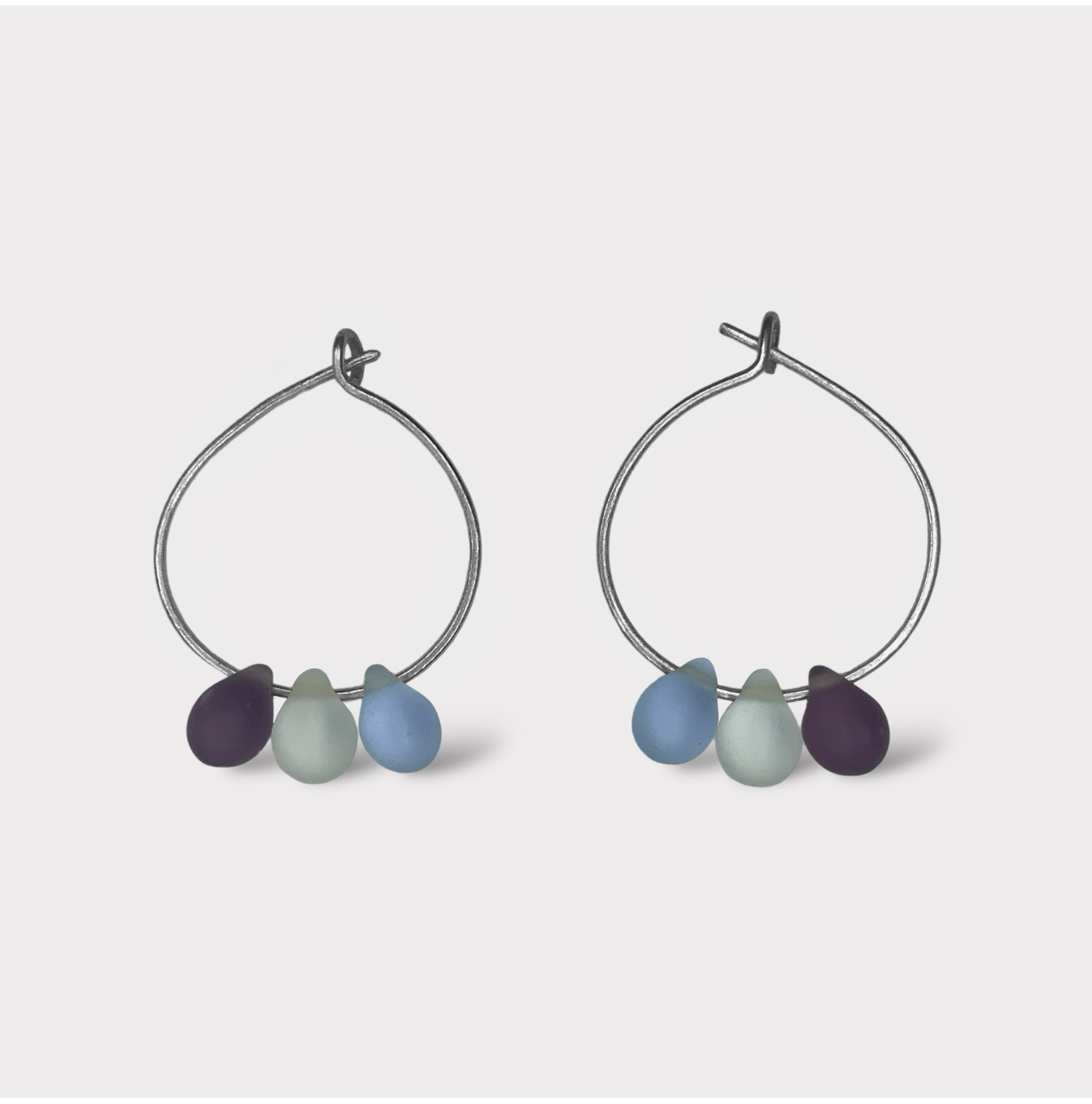 A pair of silver hoop earrings adorned with three seaglass pendants in shades of deep blue, light blue, and purple, inspired by the refreshing hues of Greek summer.