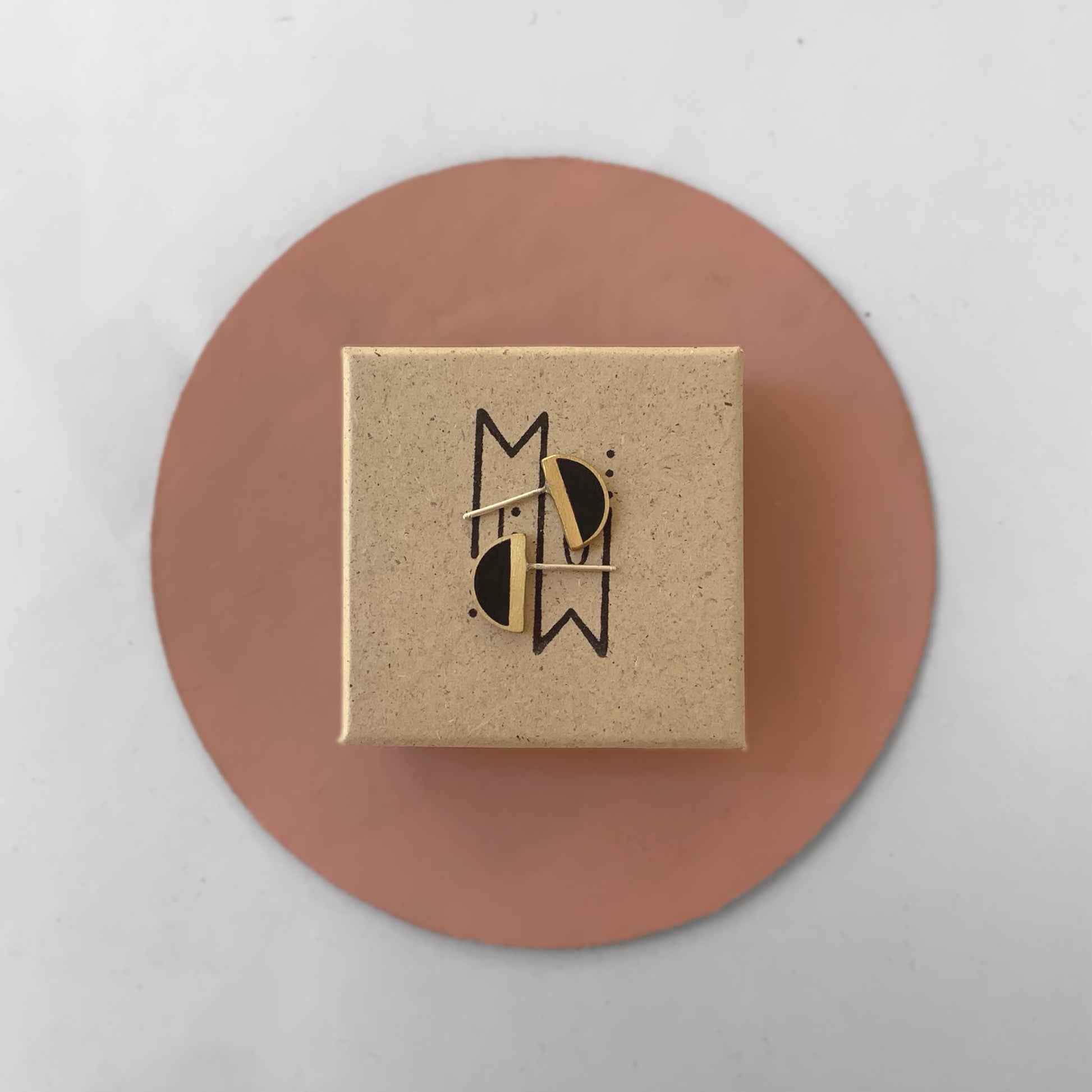 A kraft-branded gift box featuring Jewelry by Meow’s logo, adorned with brass half-cirlce earrings with black accents, styled on a pink circular accent against a white background.