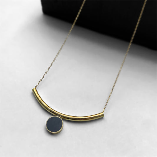A handcrafted brass necklace featuring a sleek arc-shaped pendant with a black circular charm, suspended on a delicate chain, styled against a light and dark gradient background