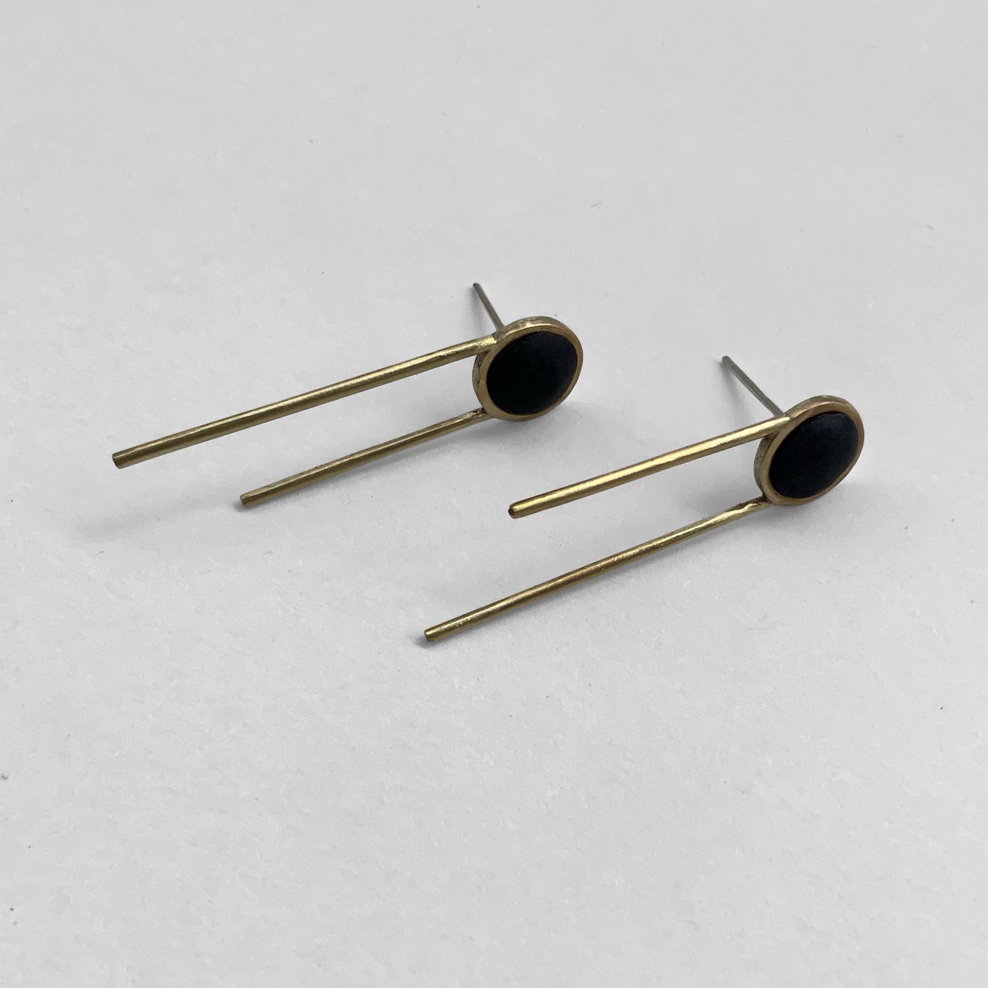 A pair of geometric stud earrings with a black circular centerpiece framed in brass, complemented by two sleek brass rods, photographed on a light gray background.

