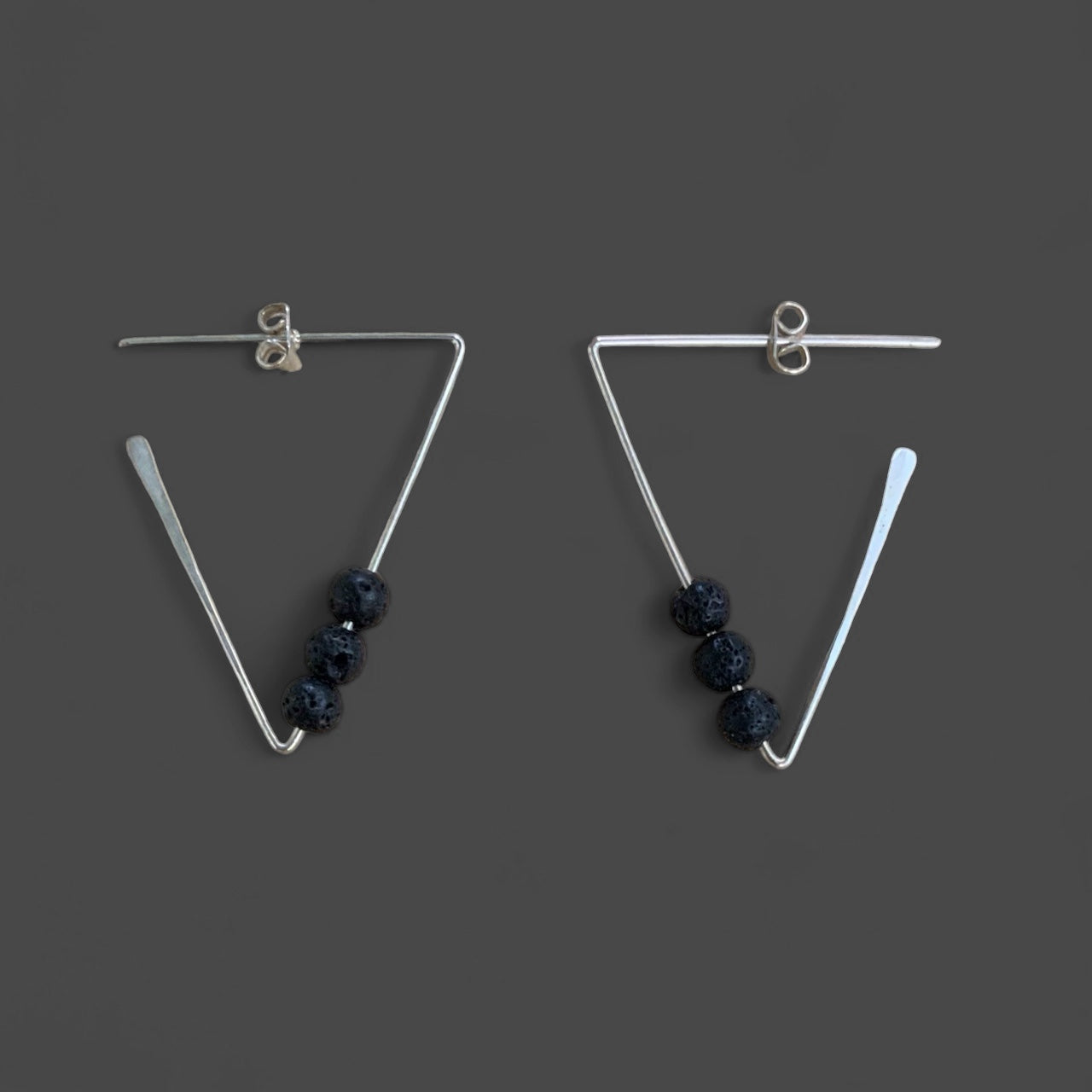 A pair of handcrafted geometric triangular silver earrings featuring three black lava beads on each side, displayed against a dark gray background. A bold and modern design with textured volcanic accents.
